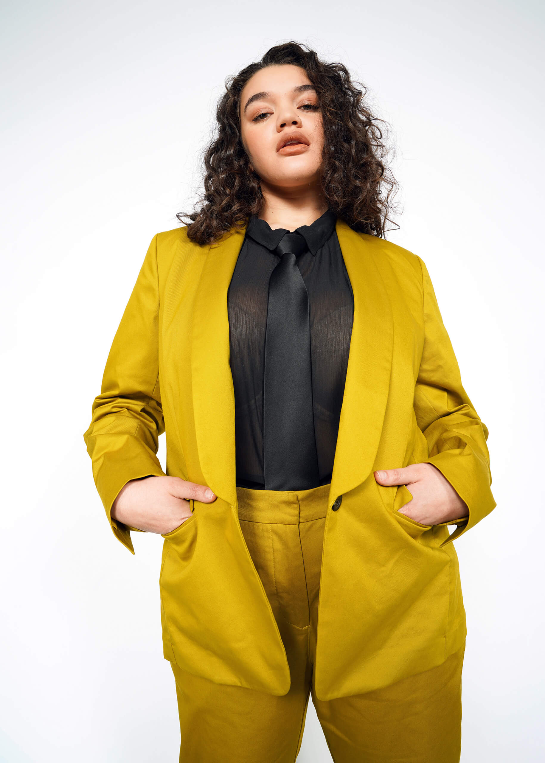A person with curly hair dons The Essential Tux Blazer in mustard yellow, paired with a black shirt and tie. They stand confidently, hands in pockets, against a plain light background.