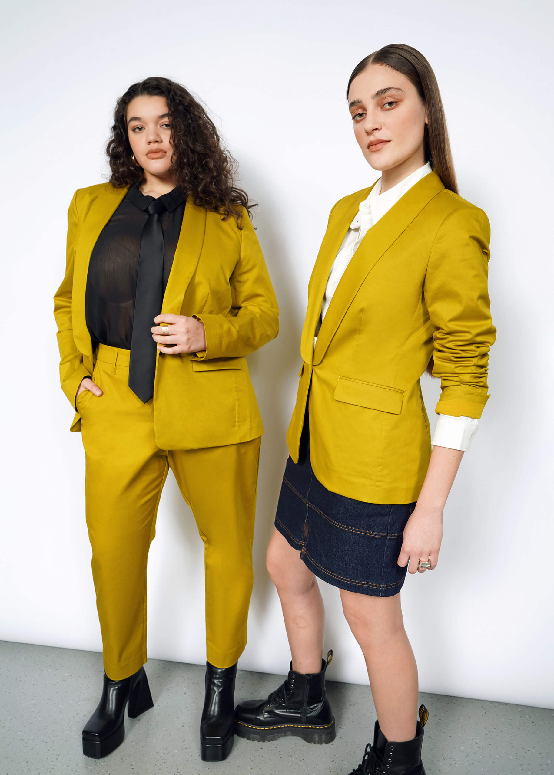 Two individuals model mustard-colored suits with tailored fits. One wears pants and a black tie, the other a skirt with a white shirt. Both sport black boots against a white wall, featuring The Essential Tux Blazer made from stretch fabric for comfort and style.