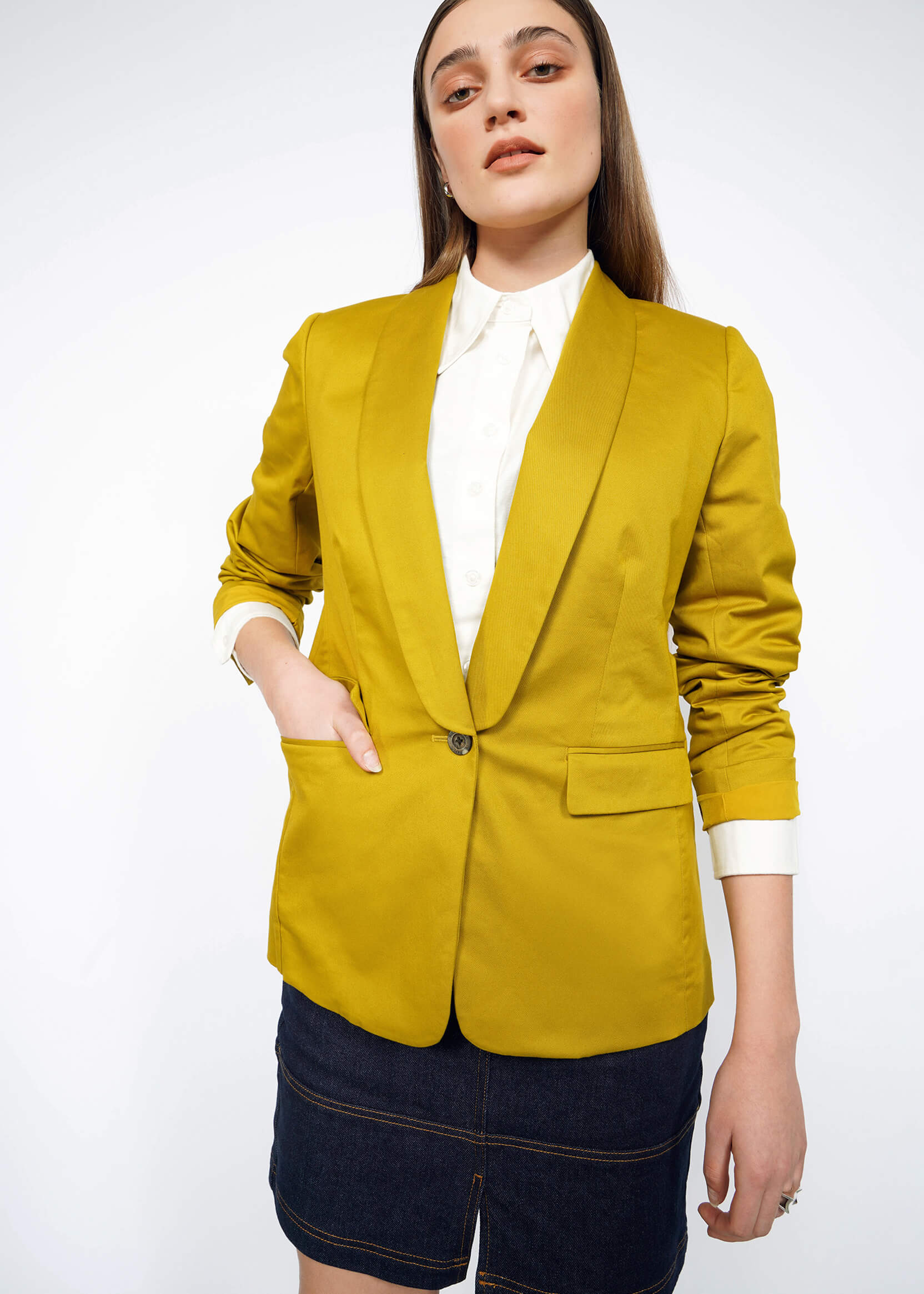 A person with long hair stands confidently in The Essential Tux Blazer, a mustard yellow tailored staple, over a white shirt and dark denim skirt against a plain background. One hand rests in their pocket while the other is relaxed by their side.