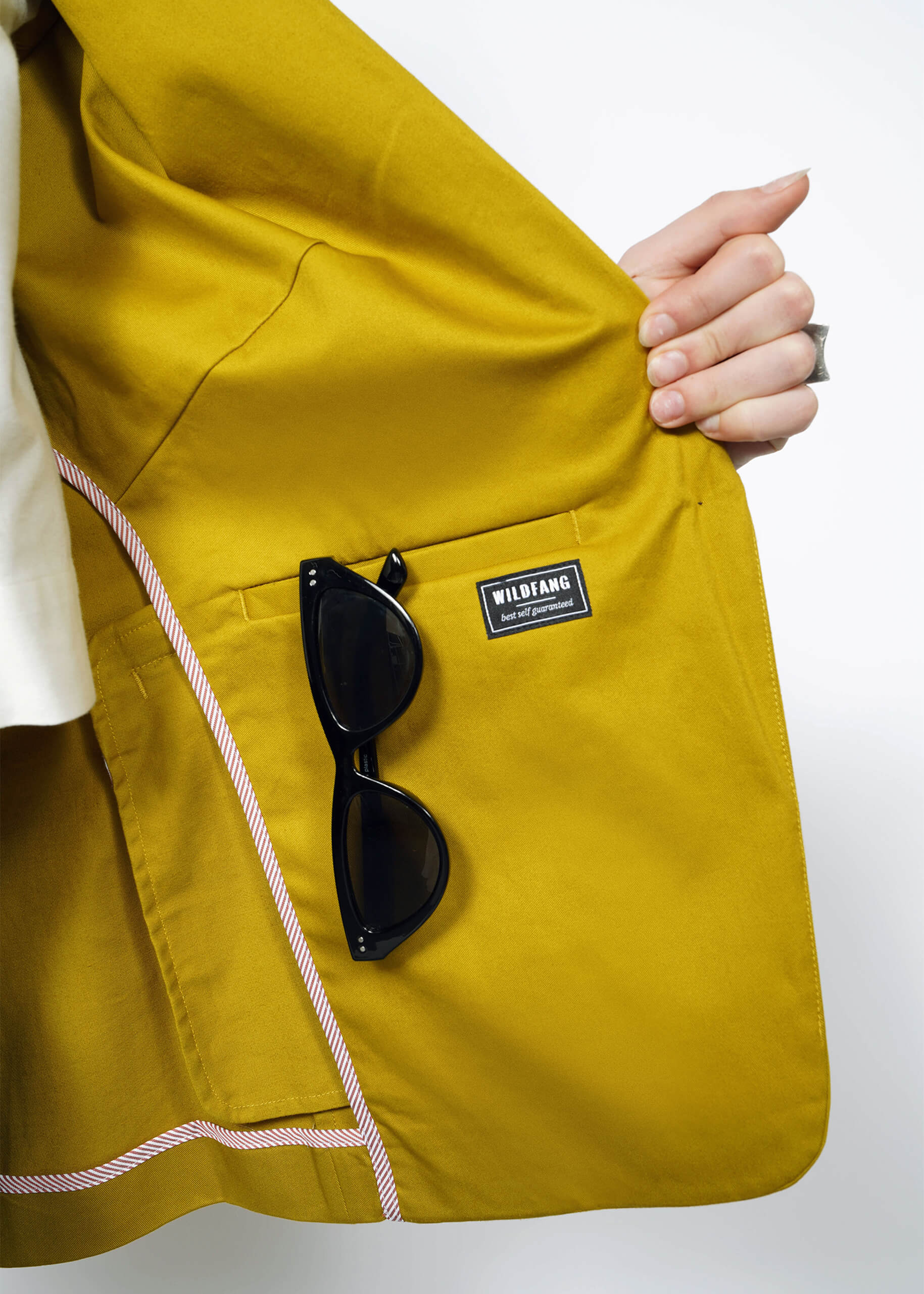 A mustard-colored jacket, The Essential Tux Blazer, has a tailored fit and pink and white striped lining. In an interior pocket with a WILDFANG label, black sunglasses are stored.