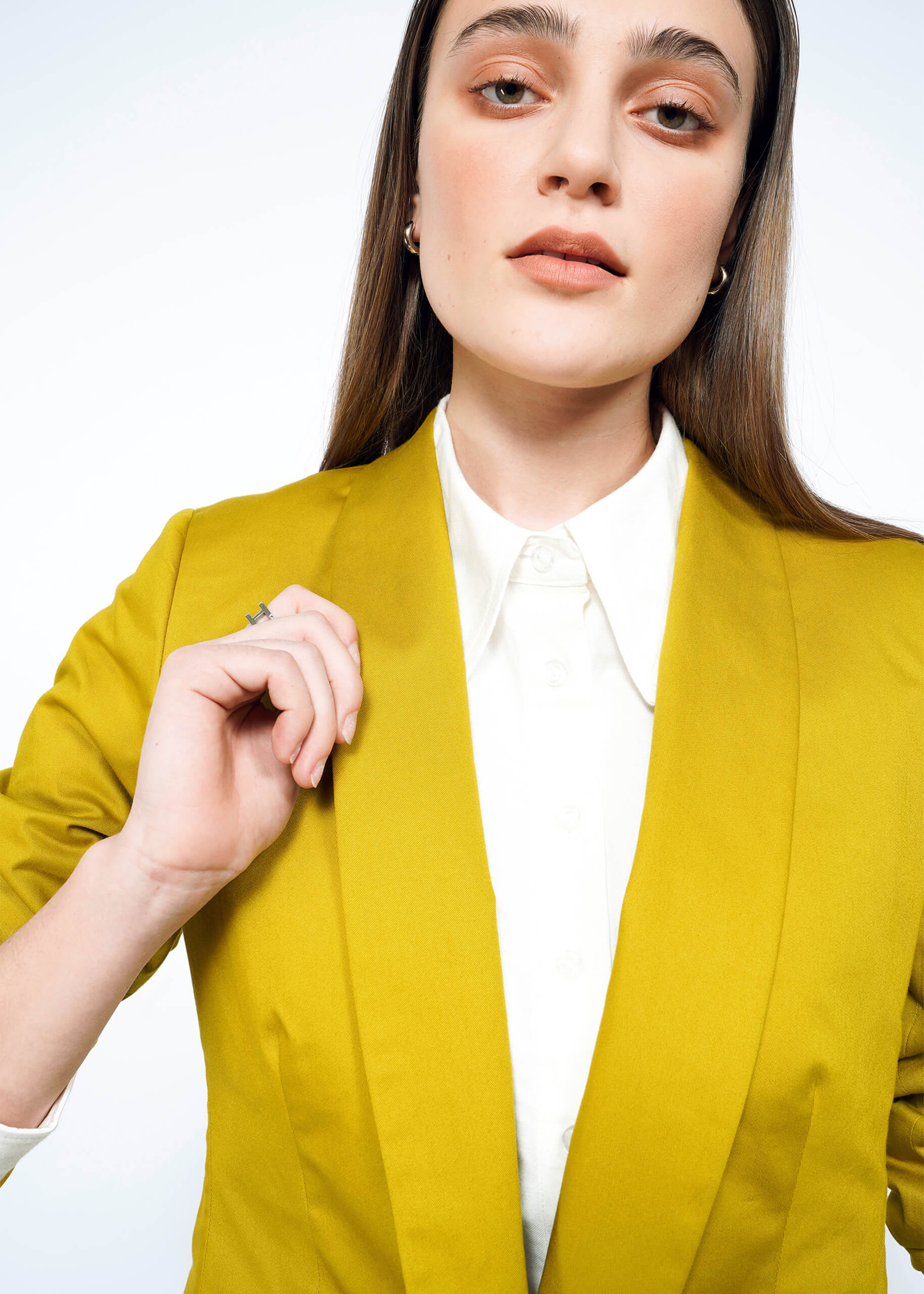 A person with long brown hair wears The Essential Tux Blazer in mustard yellow and a tailored fit over a white button-down shirt. With a neutral expression, they gently hold their lapel against a light, neutral backdrop.