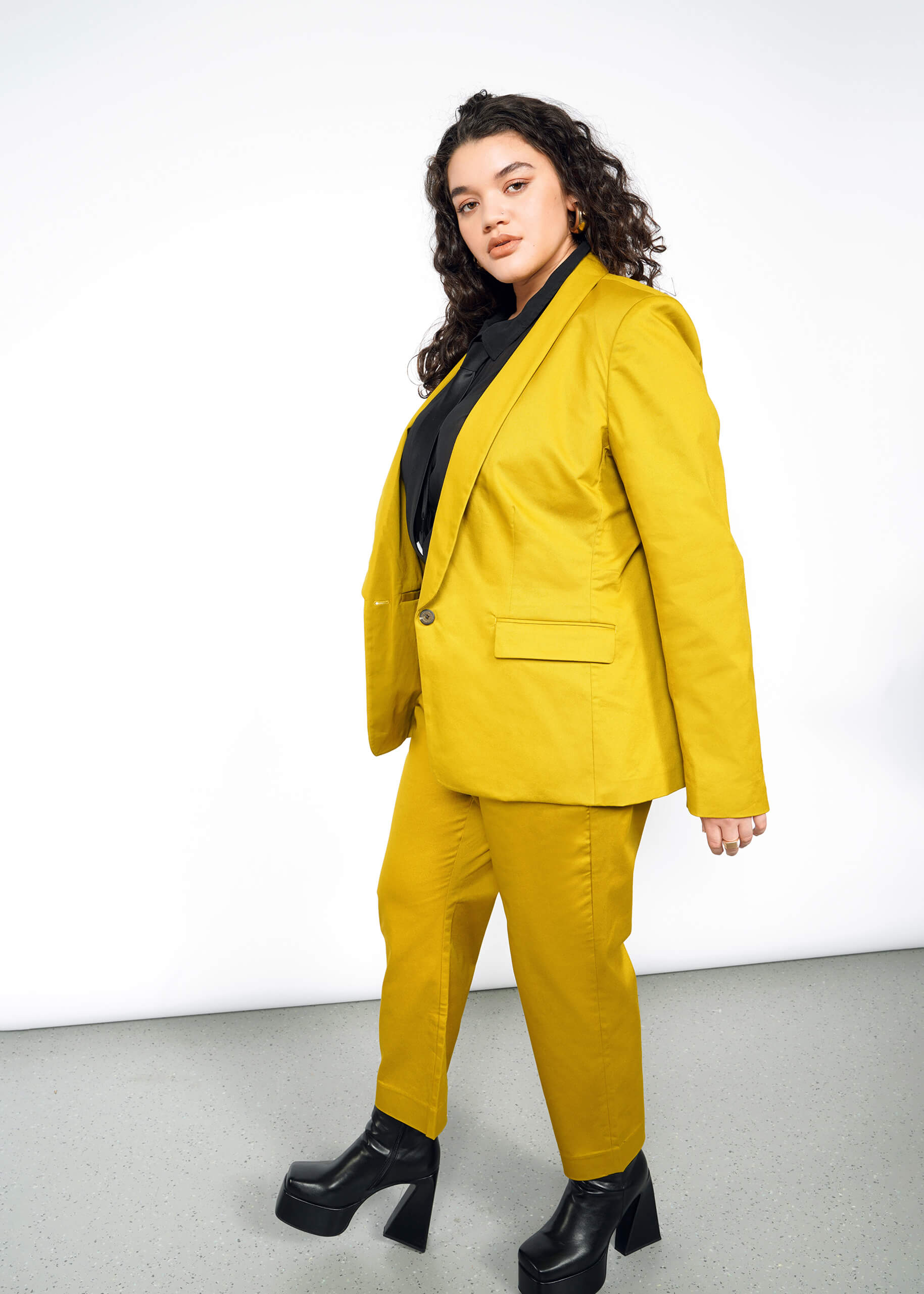 Wearing The Essential Tux Blazer in bright yellow with a tailored fit, a person with long curly hair pairs it stylishly with a black shirt and chunky platform shoes, exuding confidence against a plain white background.