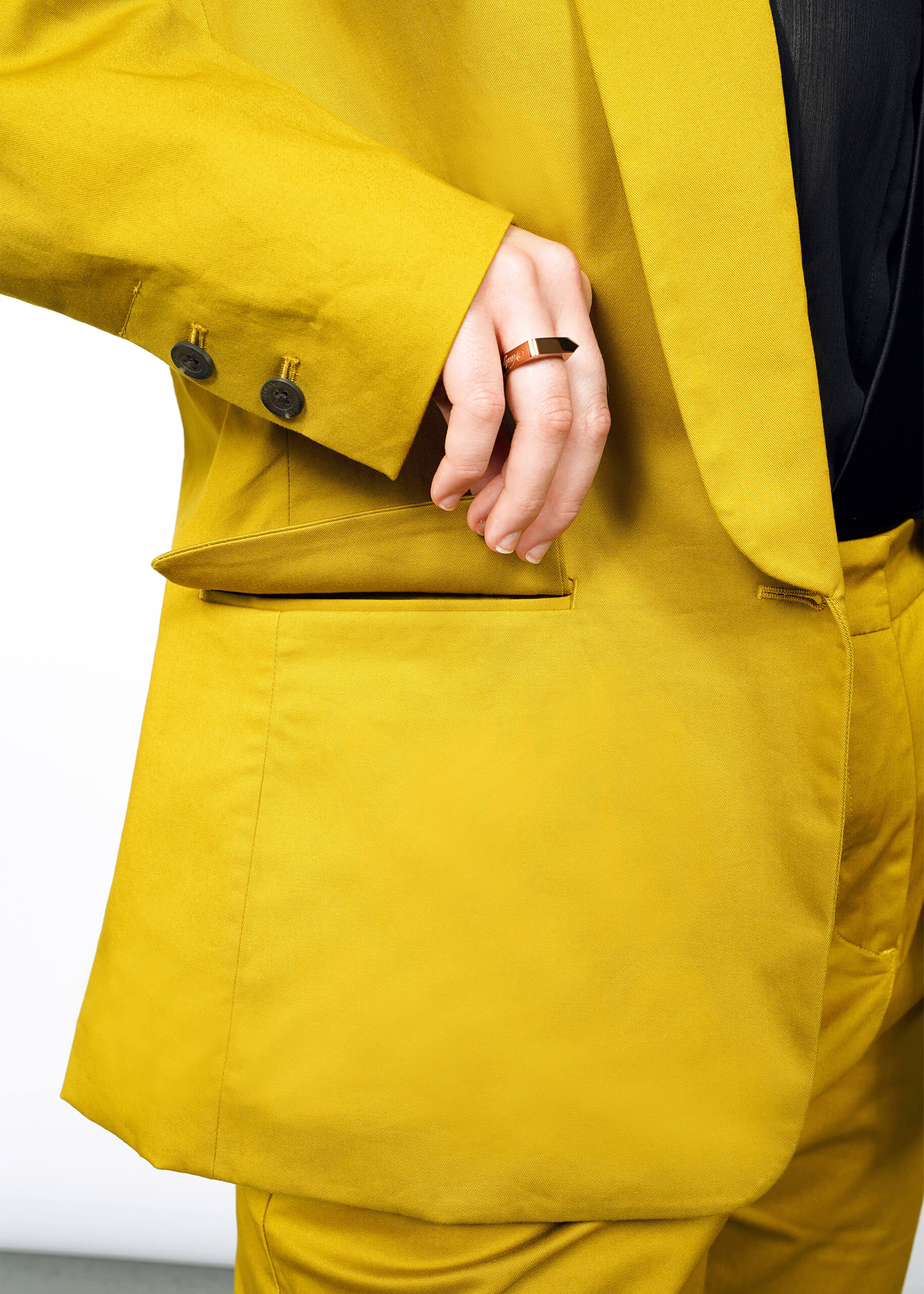 Wearing The Essential Tux Blazer in vibrant yellow with black buttons, a person places a hand adorned with a black ring into the pocket. The tailored jacket contrasts sharply against the black shirt beneath, set against a white backdrop.