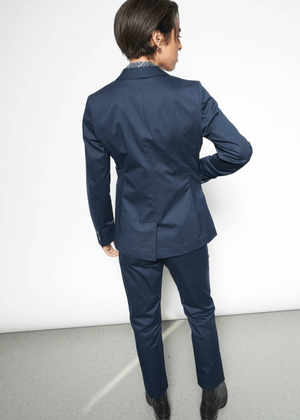 S= A person with short hair, seen from behind, is wearing a dark blue The Essential Tux Blazer in a tailored fit. Their right hand rests on their hip as they confidently pose against a plain white background.
