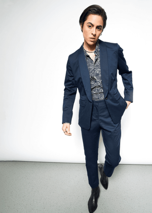 S=A person with short, dark hair confidently poses against a light grey backdrop wearing The Essential Tux Blazer in dark blue over a patterned shirt. Standing on one leg with a hand in their pocket, the tailored fit and breathable fabric exude effortless elegance.