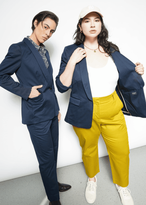 S=Two individuals flaunt their style in The Essential Tux Blazer. On the left, a tailored blue outfit paired with a patterned scarf complements the blazer, while on the right, its worn over a white shirt and yellow pants for contrast. Their ensembles pop against the crisp white backdrop.