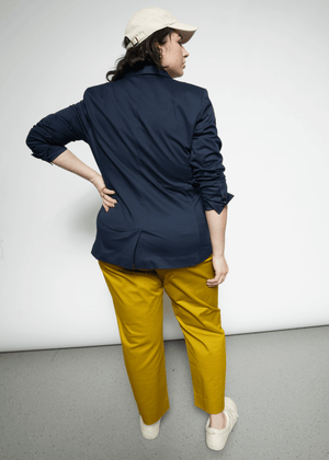 1X=Wearing The Essential Tux Blazer and mustard pants, a person stands with their back to the camera, hand on hip. A beige cap and light shoes complete the tailored look, presenting a stylish silhouette against the plain white backdrop.
