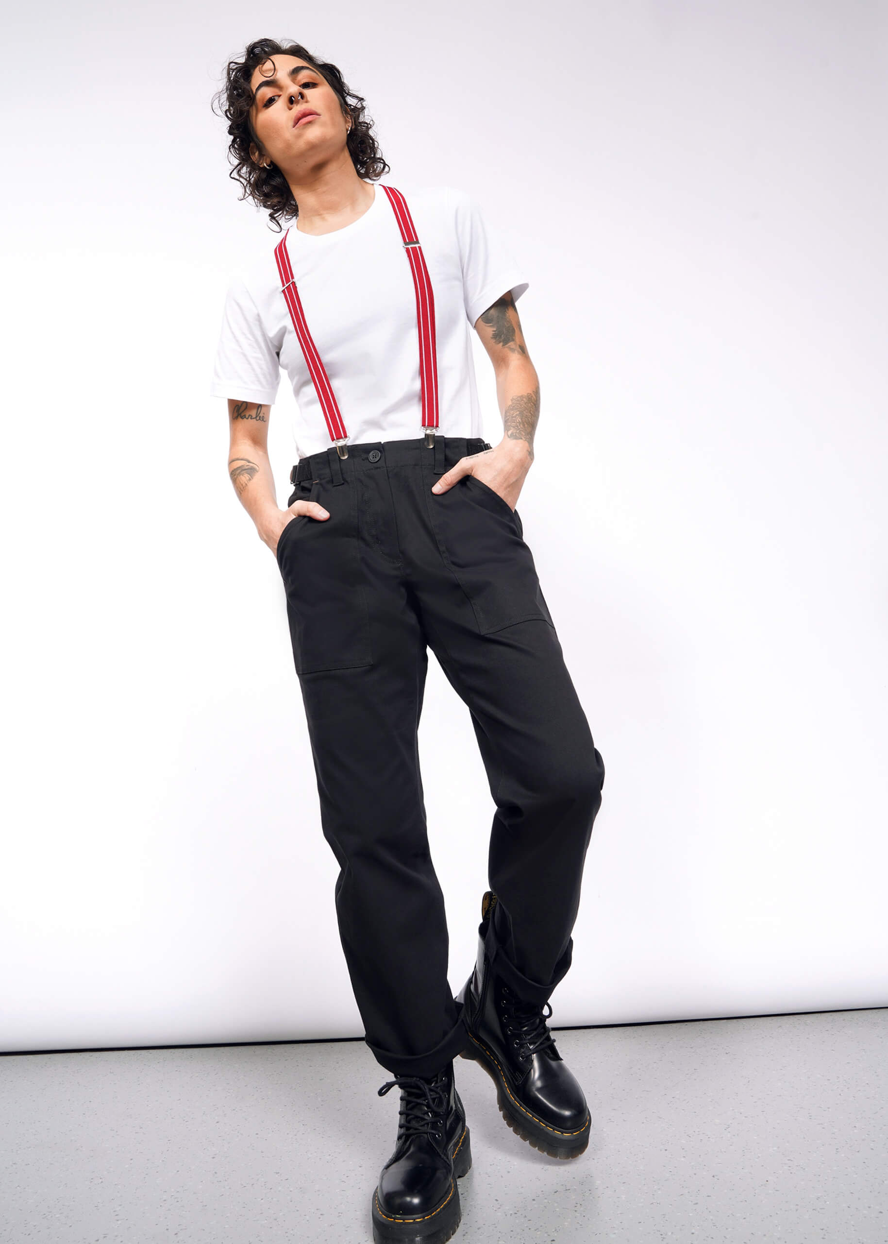 A person in a white t-shirt and black The Essential Utility Pant, featuring comfortable stretch cotton, stands confidently against a plain backdrop. Sporting red suspenders, black boots, and curly hair, they have hands in pockets with a relaxed workwear style posture.