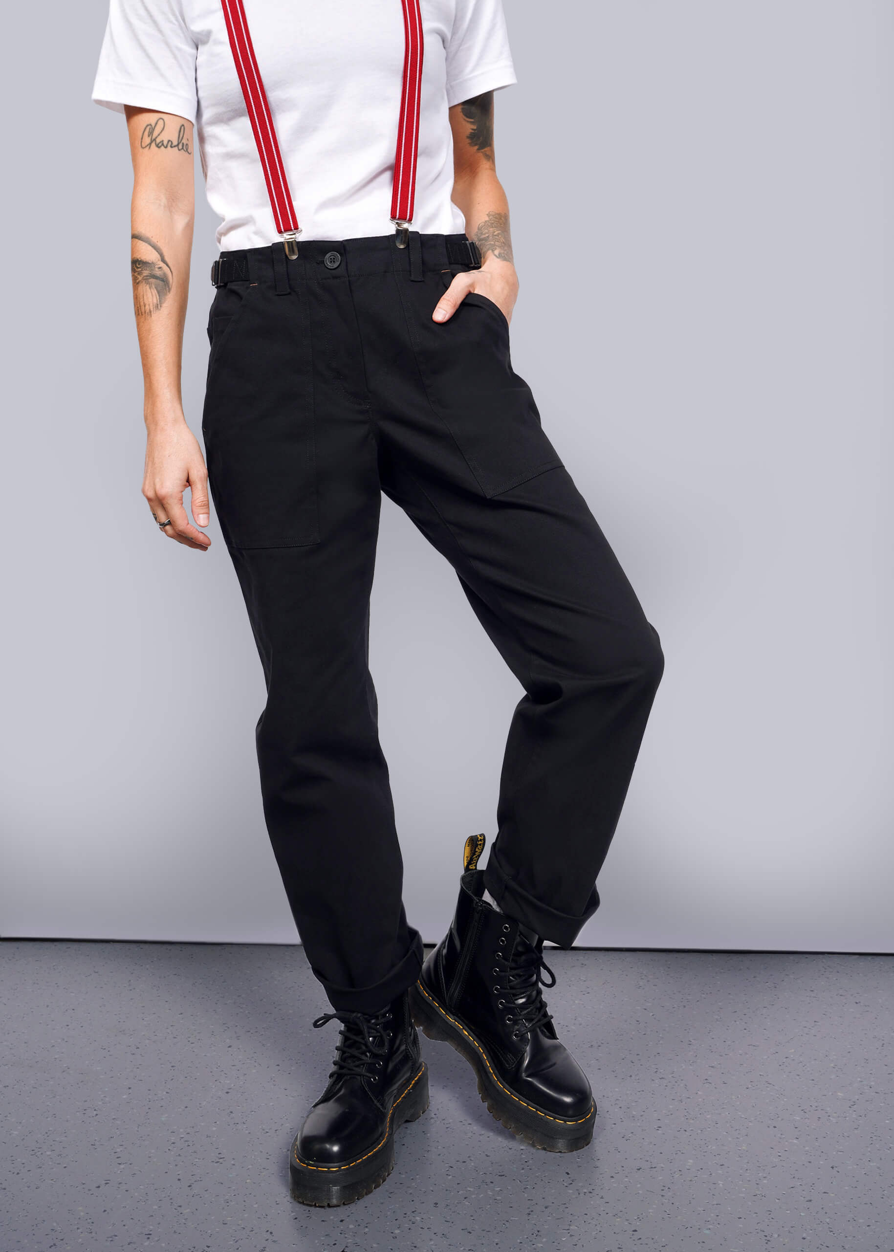 The person embodies a workwear style in black Essential Utility Pants paired with red suspenders and solid black boots. Against a plain background, their right arm rests on their hip, showcasing tattoos on both arms, while the other hand casually hangs by their side.