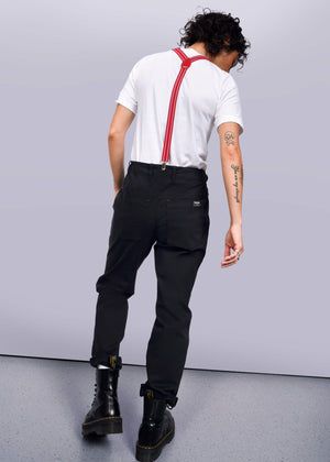 S=Facing away, the person wears a white t-shirt and black boots. Curly hair complements red suspenders over The Essential Utility Pant for a workwear look. Tattoos on their left arm add personal flair to the stretch cotton ensemble.