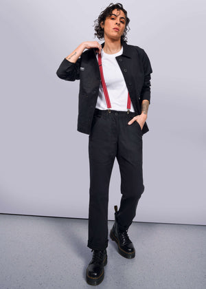 S=A person with curly hair stands confidently in a black jacket, The Essential Utility Pant, and black boots. They sport red suspenders over a stretch cotton white t-shirt, with one hand in their pocket against a simple light-colored background, embodying the workwear style.