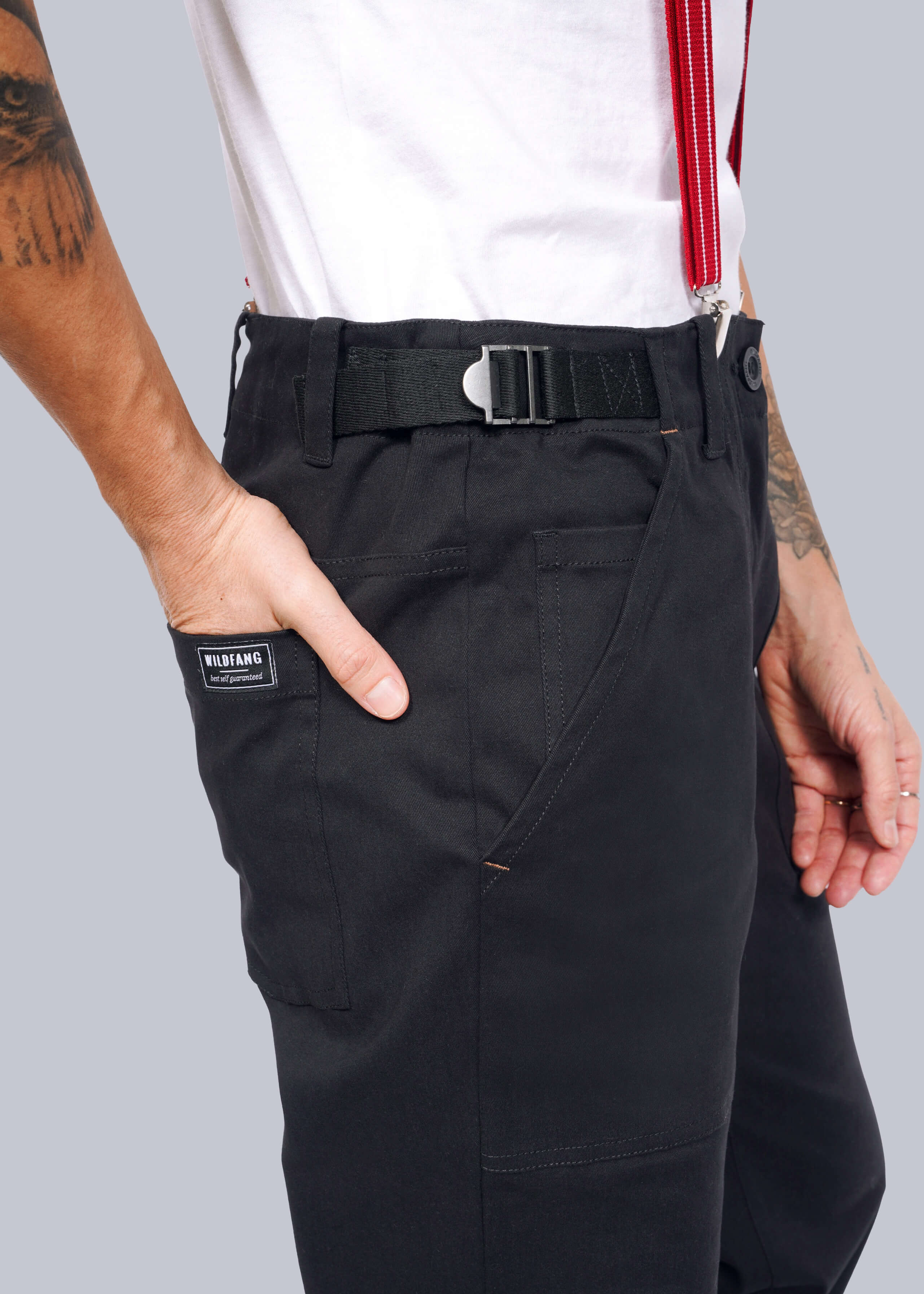 Someone wearing The Essential Utility Pant in black, featuring a sleek belt and bold red suspenders. A hand casually rests in the pocket, showcasing the label. The stretch cotton fabric pairs with a crisp white shirt for a seamless modern workwear look.