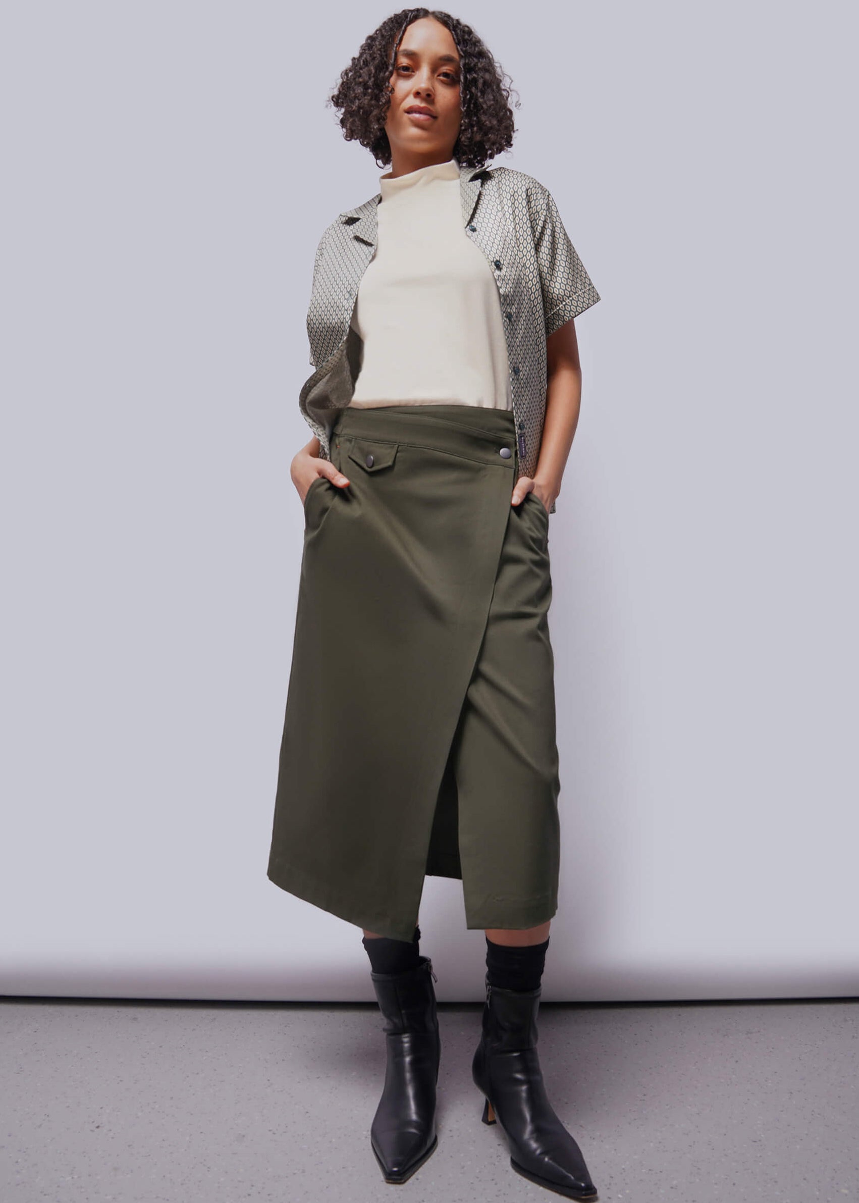 S=Person wearing a white shirt, patterned cardigan, green skirt, and black boots against a plain background