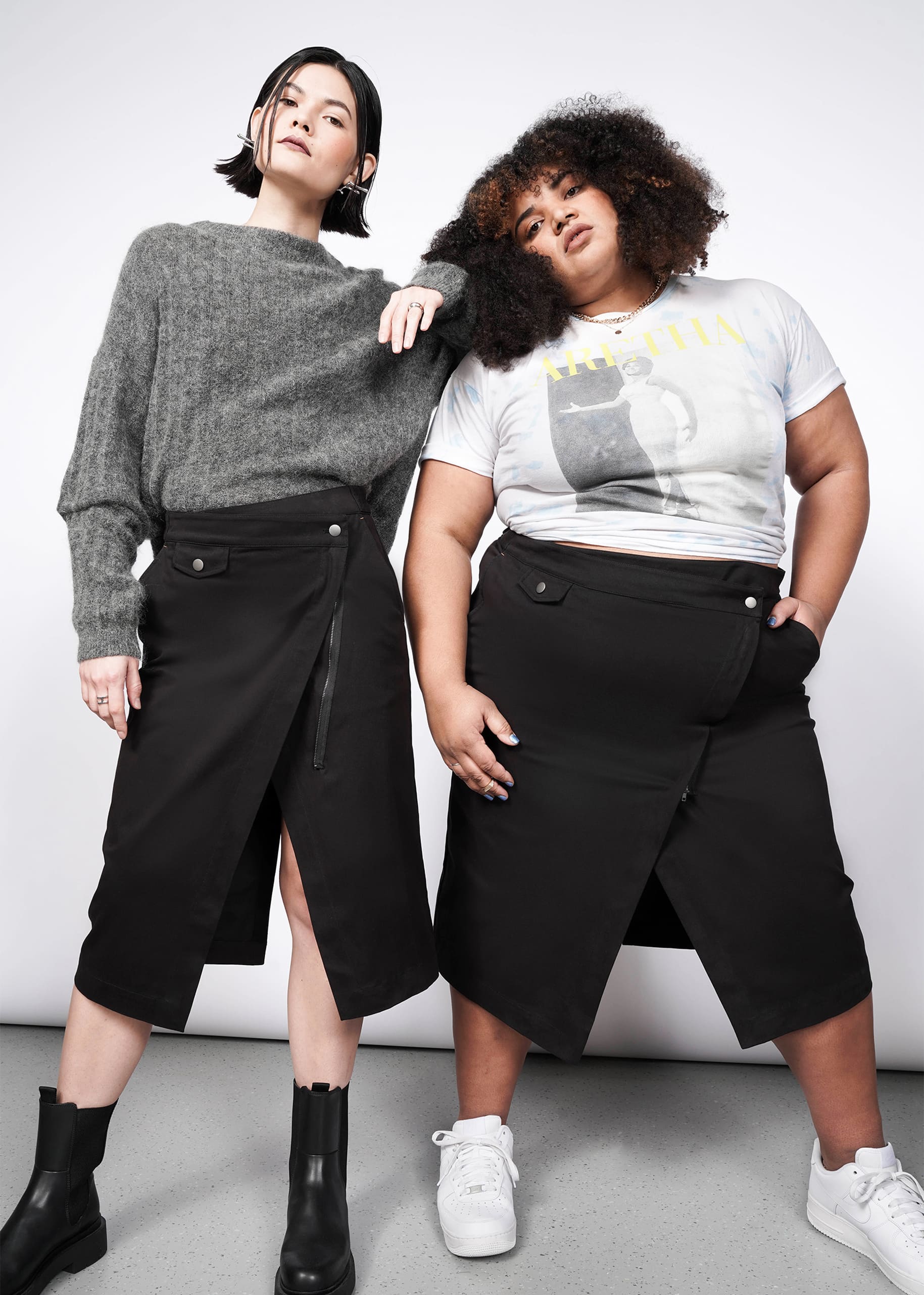 Two women showcase The Essential Utility Wrap Skirt, styled with a gray sweater and beanie or a graphic tee and sneakers. Each pairs the black midi skirt’s front slit with unique looks, standing against a plain background.