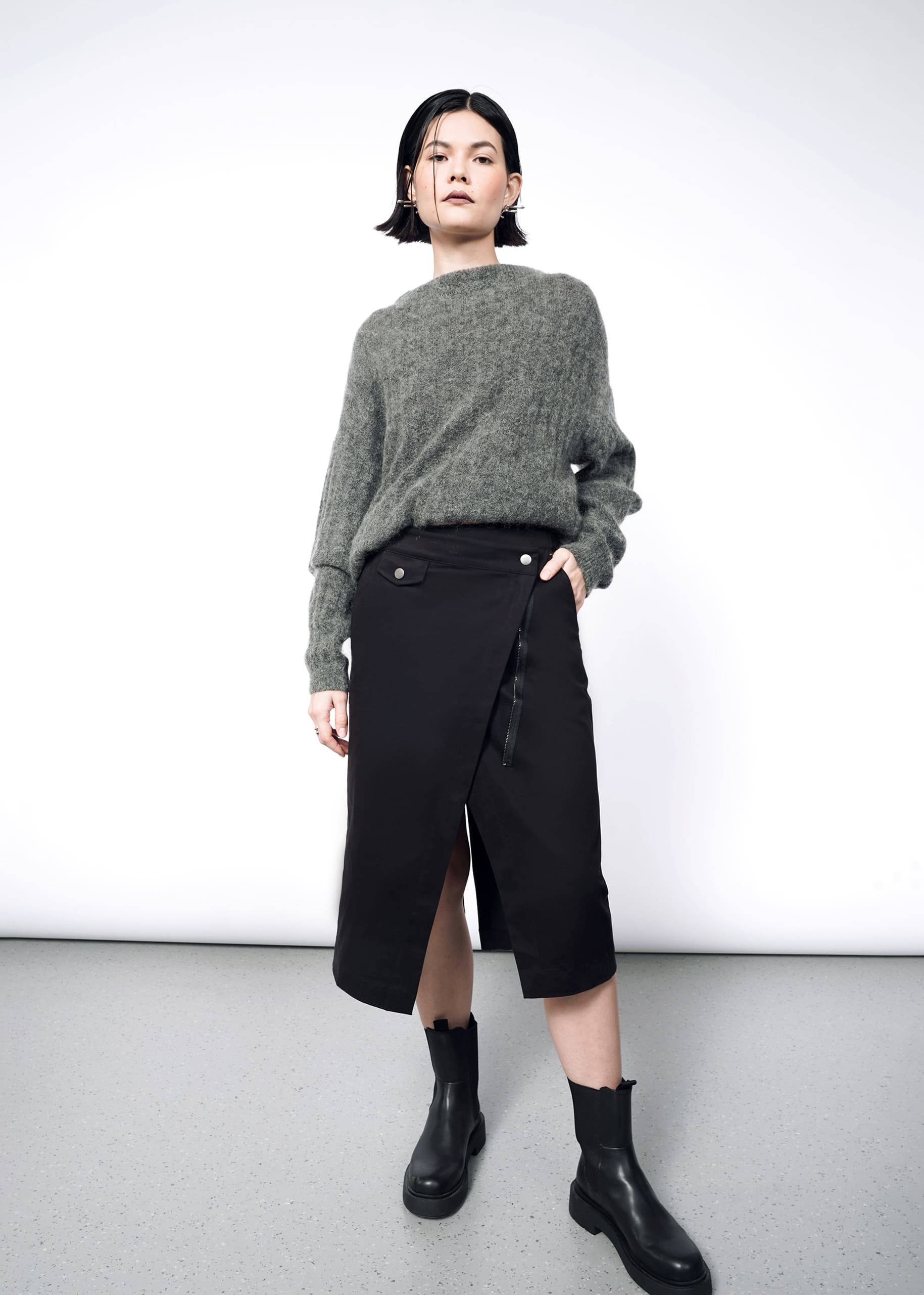 A person stands confidently against a plain white background, wearing a loose gray sweater, black ankle boots, and The Essential Utility Wrap Skirt featuring an asymmetrical waist detail and side strap.