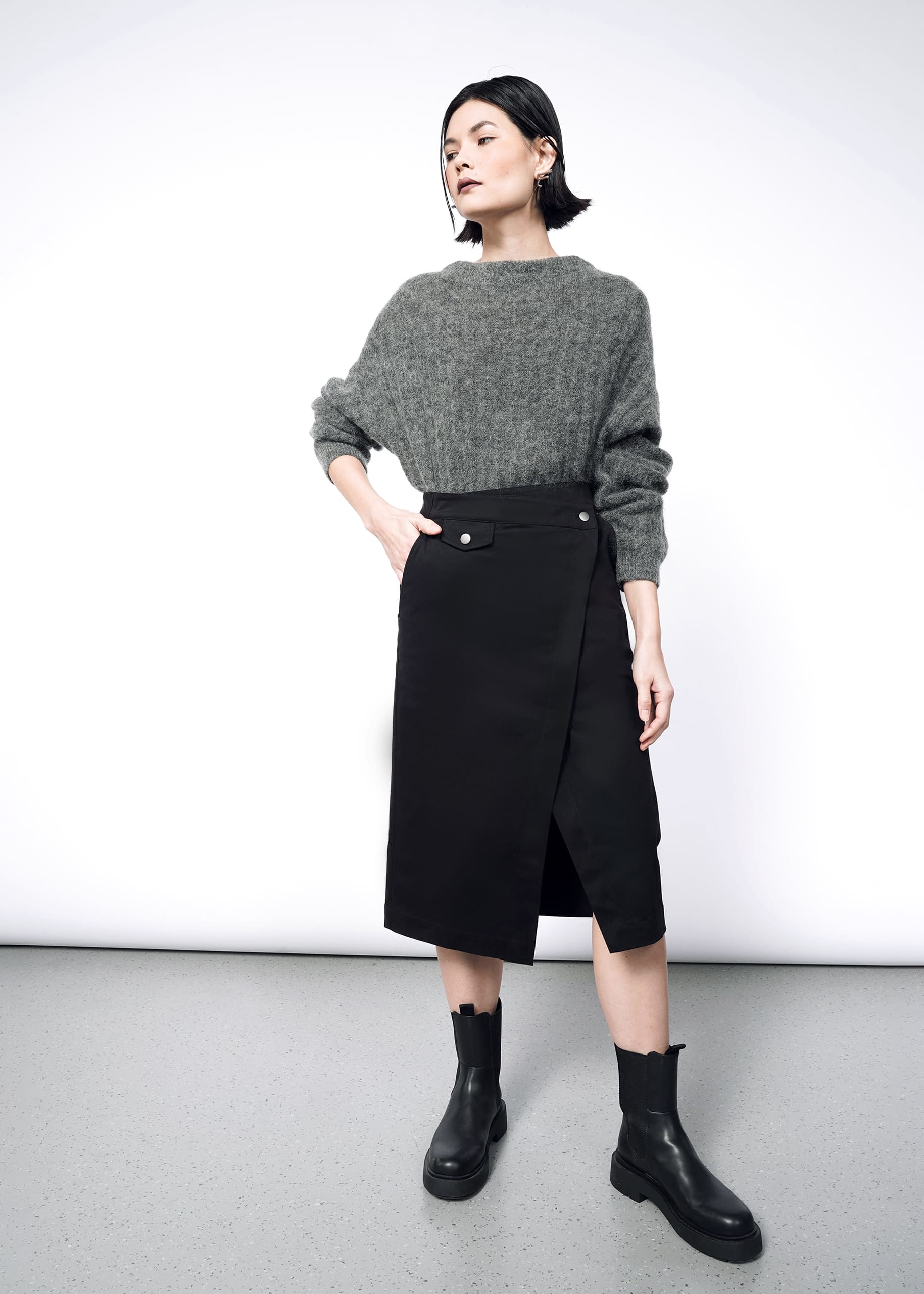 A woman poses against a white backdrop in a gray sweater and The Essential Utility Wrap Skirt—black, midi length, with buttons and an asymmetric waist. She pairs the look with black ankle boots and styles her short dark hair confidently.