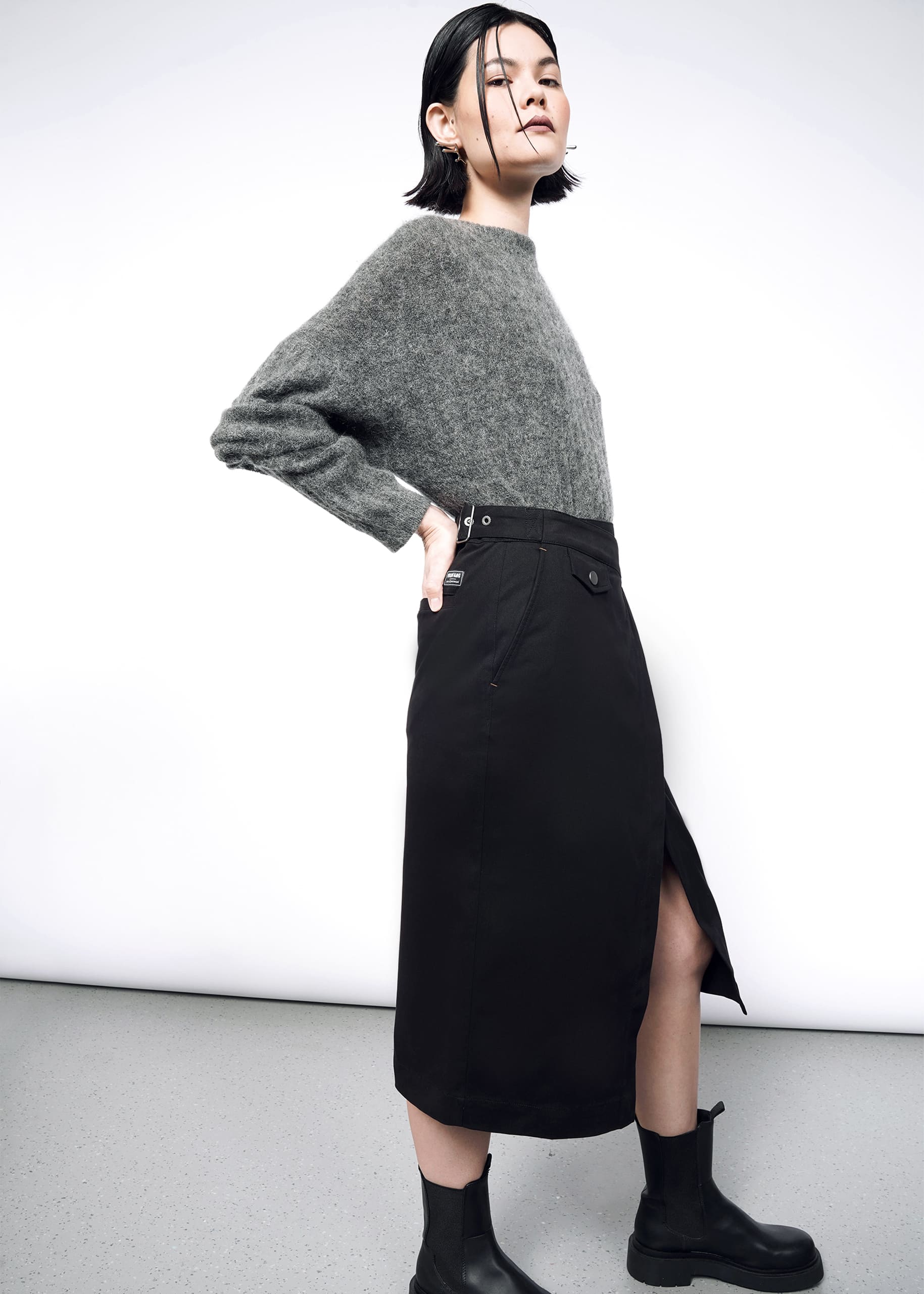 A woman stands sideways against a white background, confidently posing with one hand on her hip in a gray fuzzy sweater, black ankle boots, and The Essential Utility Wrap Skirt featuring an asymmetrical waist detail.