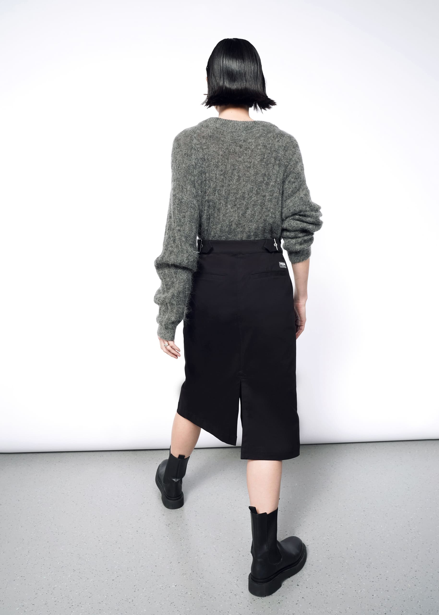 A person with short dark hair faces away, wearing a loose gray sweater with The Essential Utility Wrap Skirt—black, midi-length with back slit and adjustable waistband—and black ankle boots, against a plain white background.