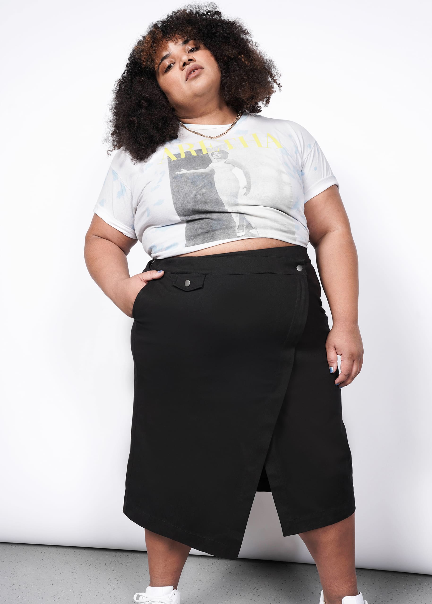 A person with curly hair wears a printed crop top, The Essential Utility Wrap Skirt in black with an asymmetrical waist detail, and white sneakers, standing against a plain white wall and looking confidently at the camera.