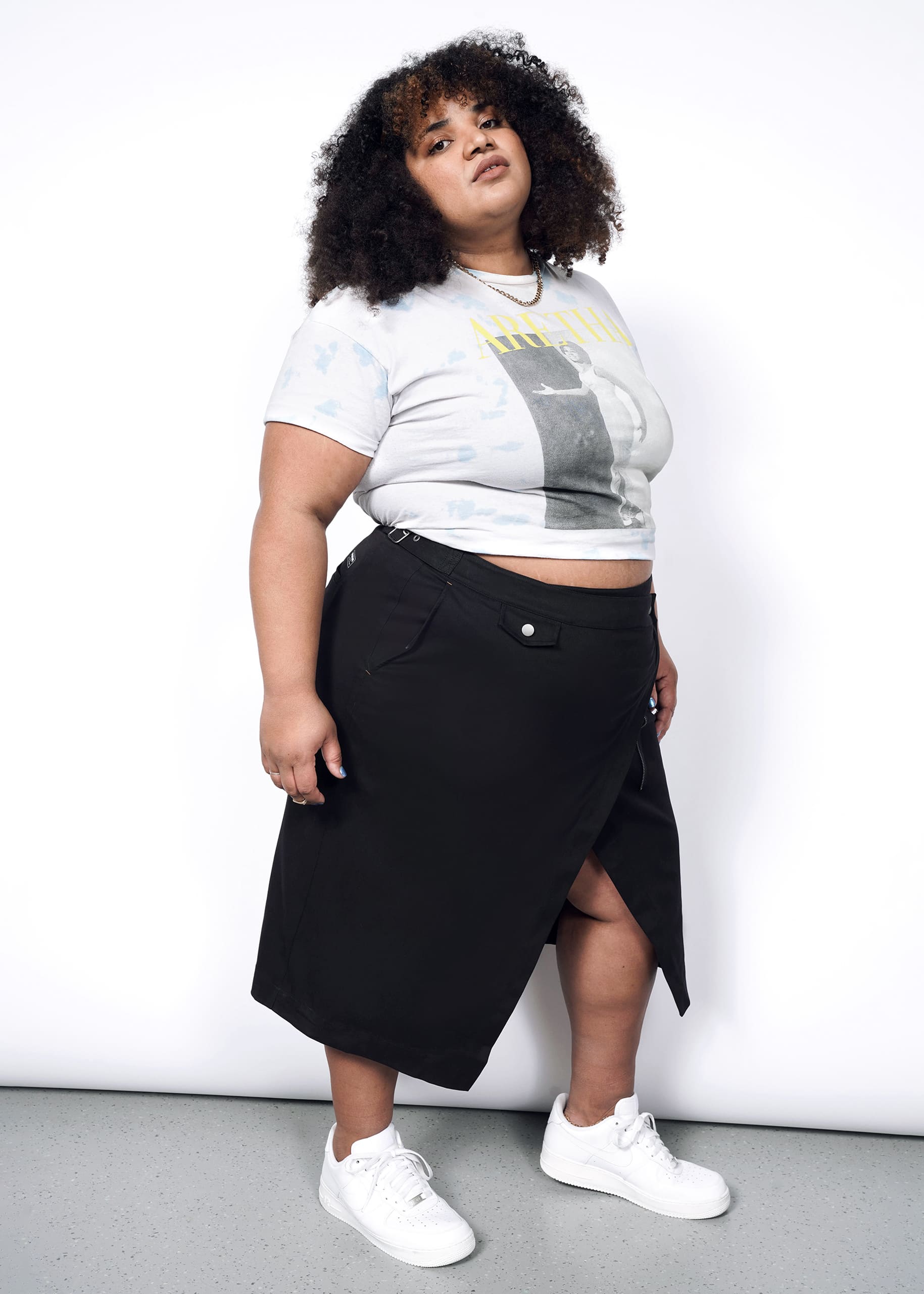A person with curly hair wears a graphic crop top, The Essential Utility Wrap Skirt in black with an asymmetrical waist detail, and white sneakers, standing confidently against a plain white background.