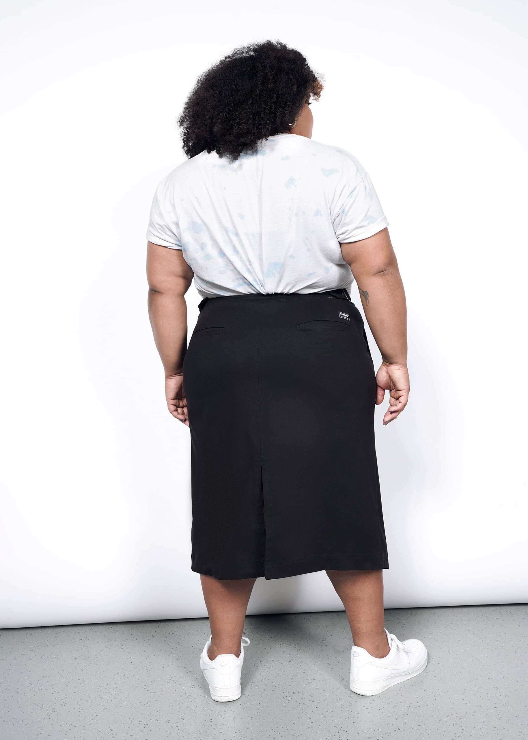 A person with curly hair faces away, wearing a light t-shirt, The Essential Utility Wrap Skirt in black with an asymmetrical waist, and white sneakers against a white background.