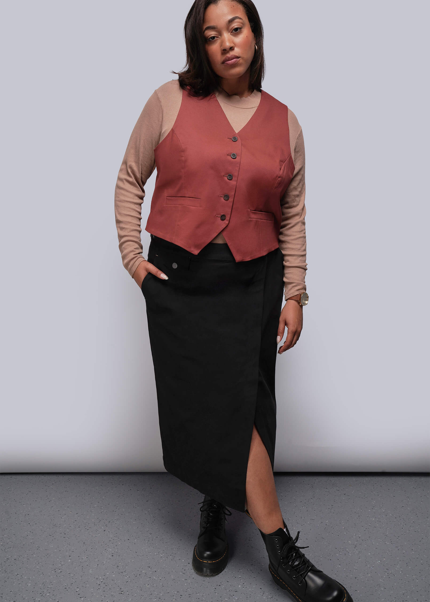 1X=Person wearing a rust-colored vest over a beige long-sleeve shirt, black skirt, and black shoes on a plain background.