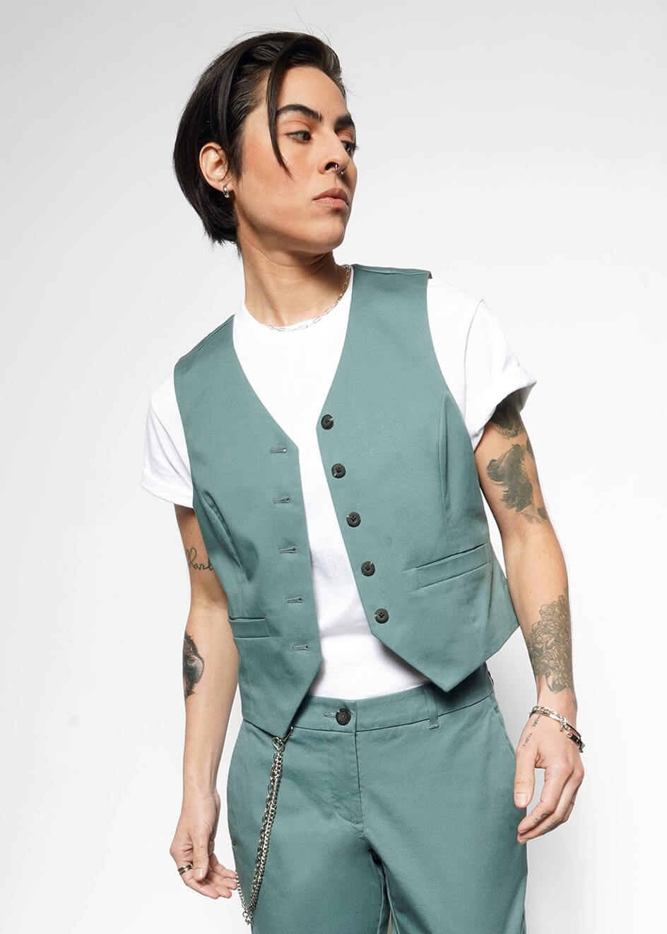 A person with short dark hair wears The Essential Vest and matching stretch cotton pants over a white t-shirt, accessorized with bracelets and a chain, standing against a plain white background.