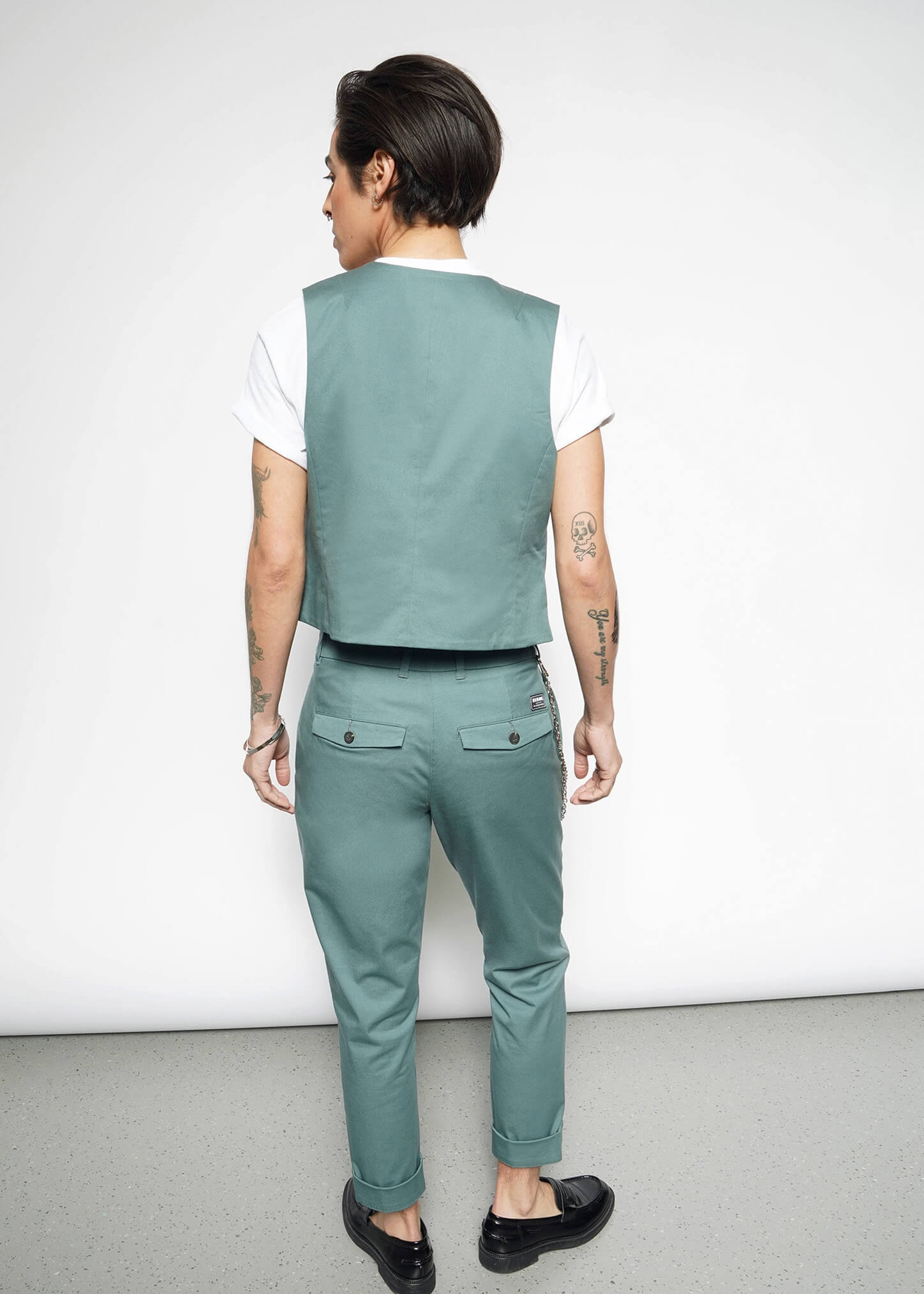A person with short dark hair, wearing The Essential Vest—tailored fit, stretch cotton, 5-button closure—with matching pants, white T-shirt, and black shoes stands facing away from the camera, displaying arm tattoos against a plain white background.