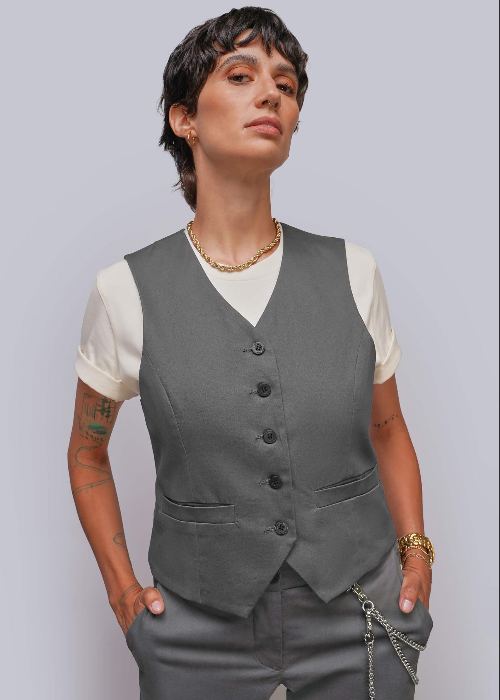 A model wearing a dark gray button-up vest over a cream t-shirt, paired with gray trousers and gold jewelry, set against a solid light gray background.