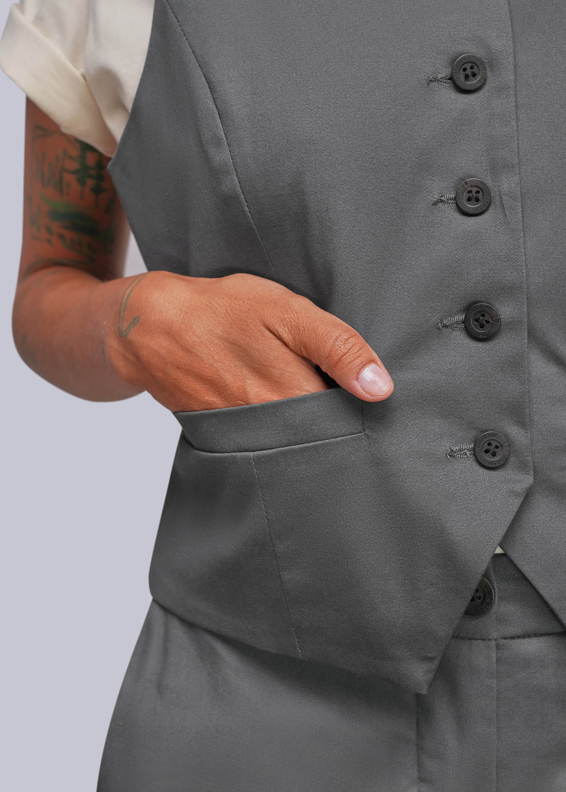 Close-up of a person wearing a gray tailored vest with dark buttons and a side pocket, with one hand inserted into the pocket. A cream short-sleeve shirt is partially visible.
