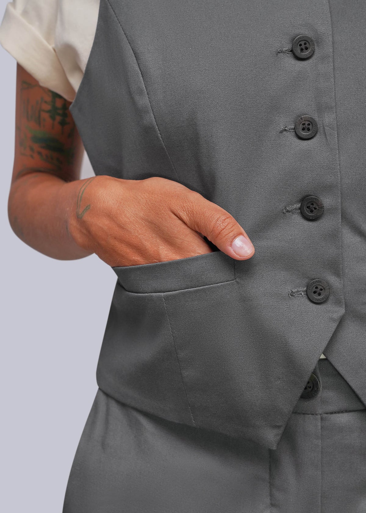 S=Close-up of a person wearing a gray tailored vest with dark buttons and a side pocket, with one hand inserted into the pocket. A cream short-sleeve shirt is partially visible.