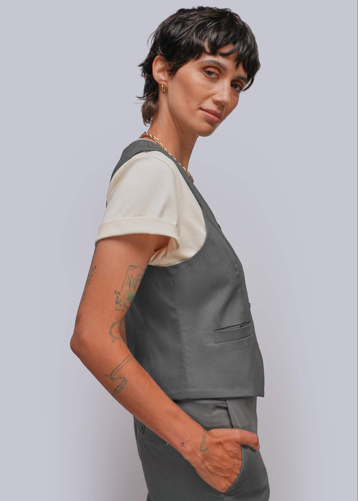 S=A model with short dark hair is shown in a side profile wearing a dark gray sleeveless vest over a cream short-sleeve t-shirt and matching gray trousers, set against a solid light gray background.