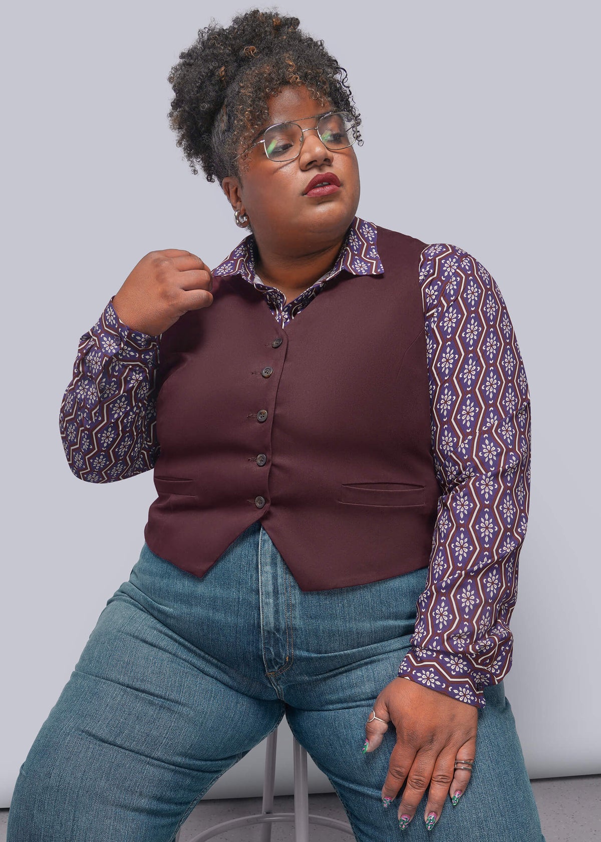 1X=A plus-size model wearing a burgundy button-up vest over a purple and white patterned long-sleeve shirt, paired with blue jeans, sitting on a stool against a light gray studio background.