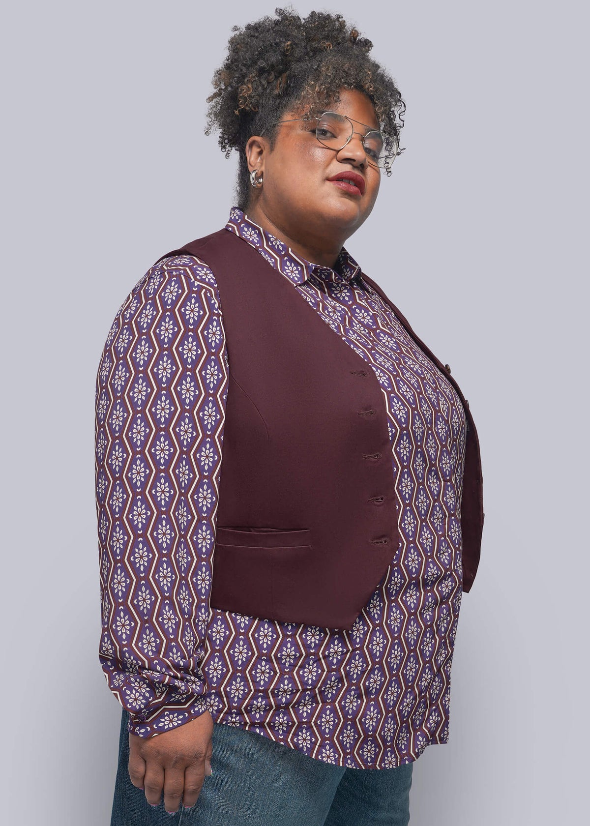 1X=A person wearing a burgundy vest over a purple and white patterned button-up shirt, paired with blue jeans, photographed against a solid light gray studio background.