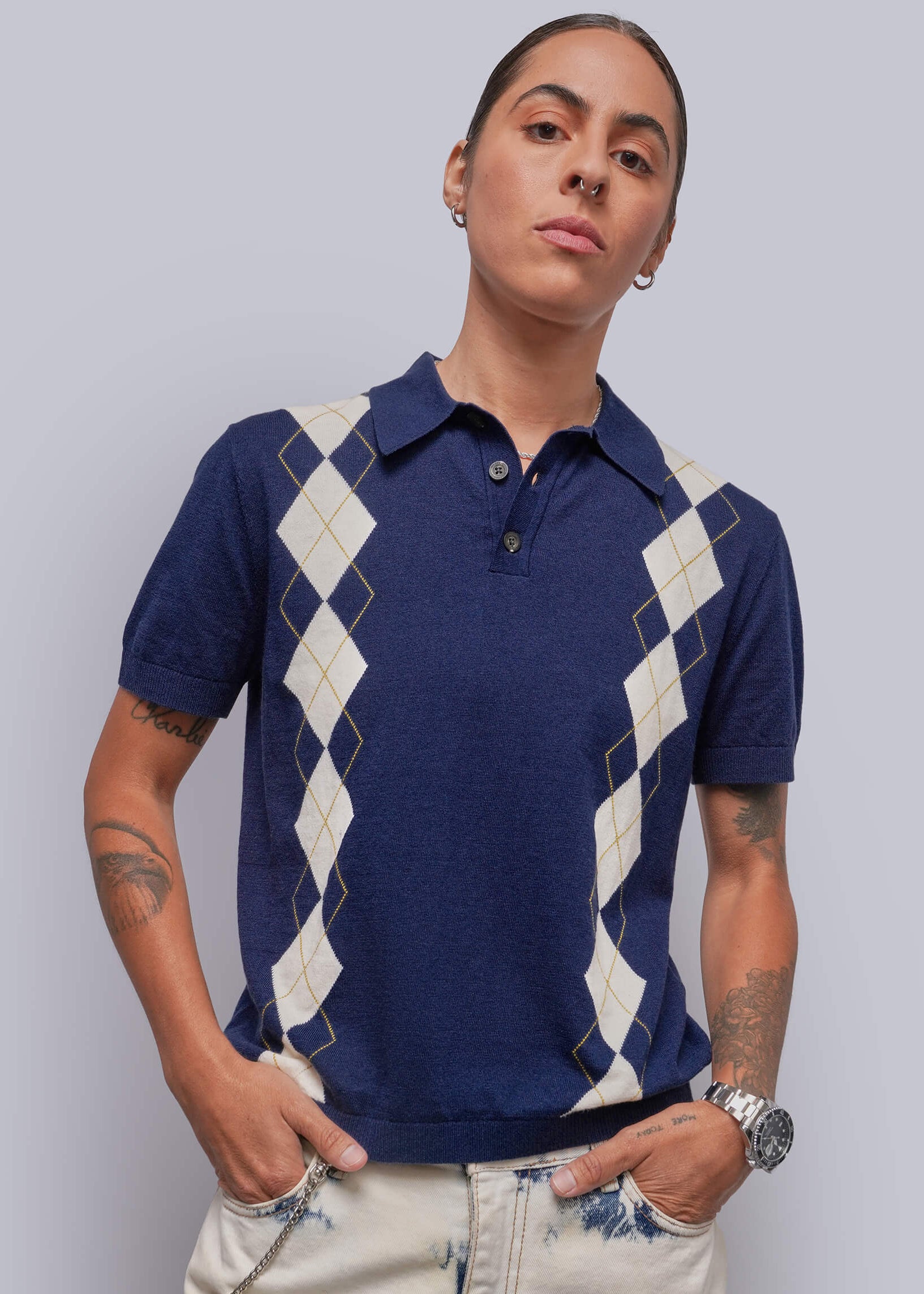 S=A model wearing a navy blue short-sleeve polo shirt with cream argyle diamond patterns on the chest and sleeves, paired with light beige jeans, against a solid light gray background.