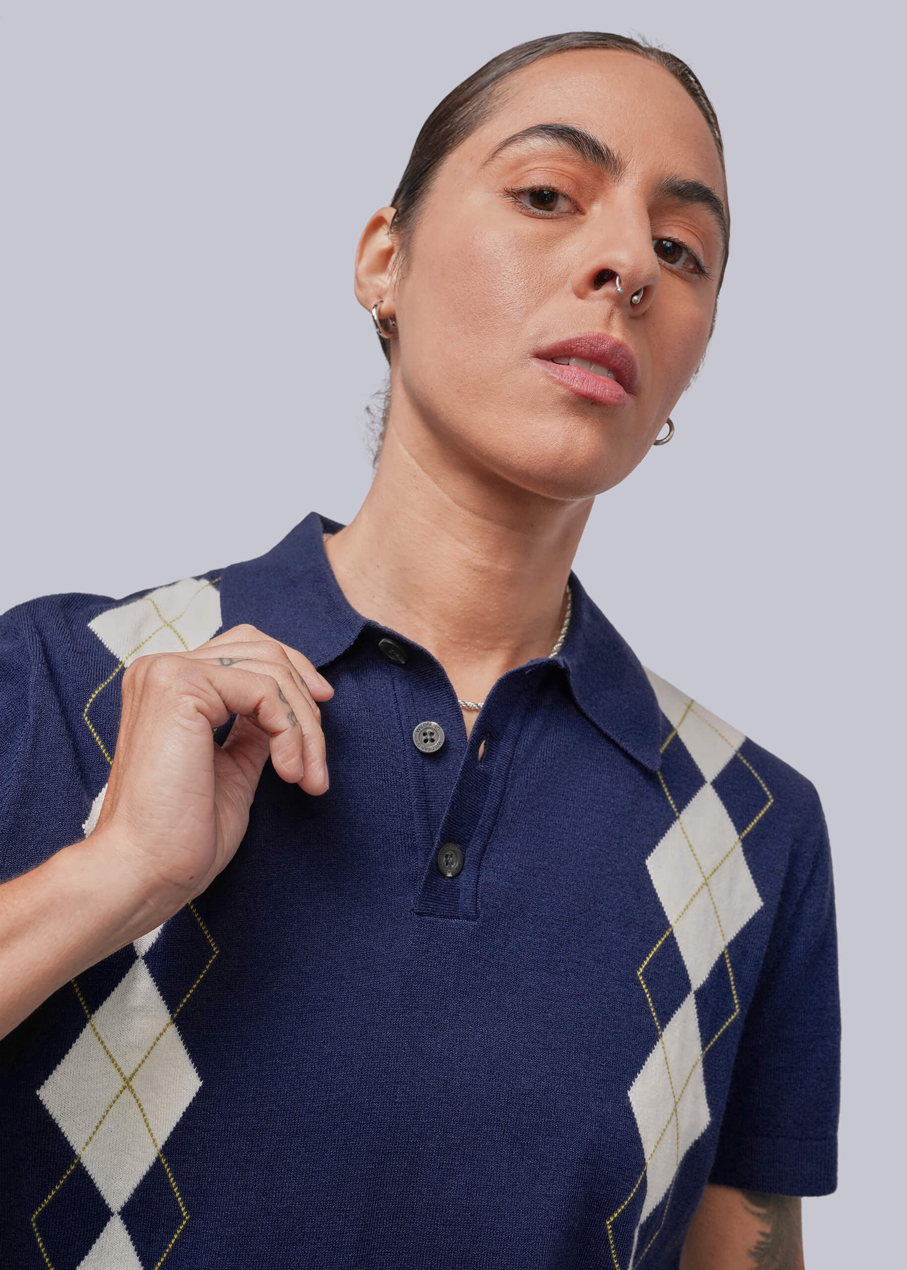 S=A close-up of a person wearing a navy blue short-sleeve polo shirt with cream and gold argyle diamond patterns on the shoulders and sides, against a solid light gray background.