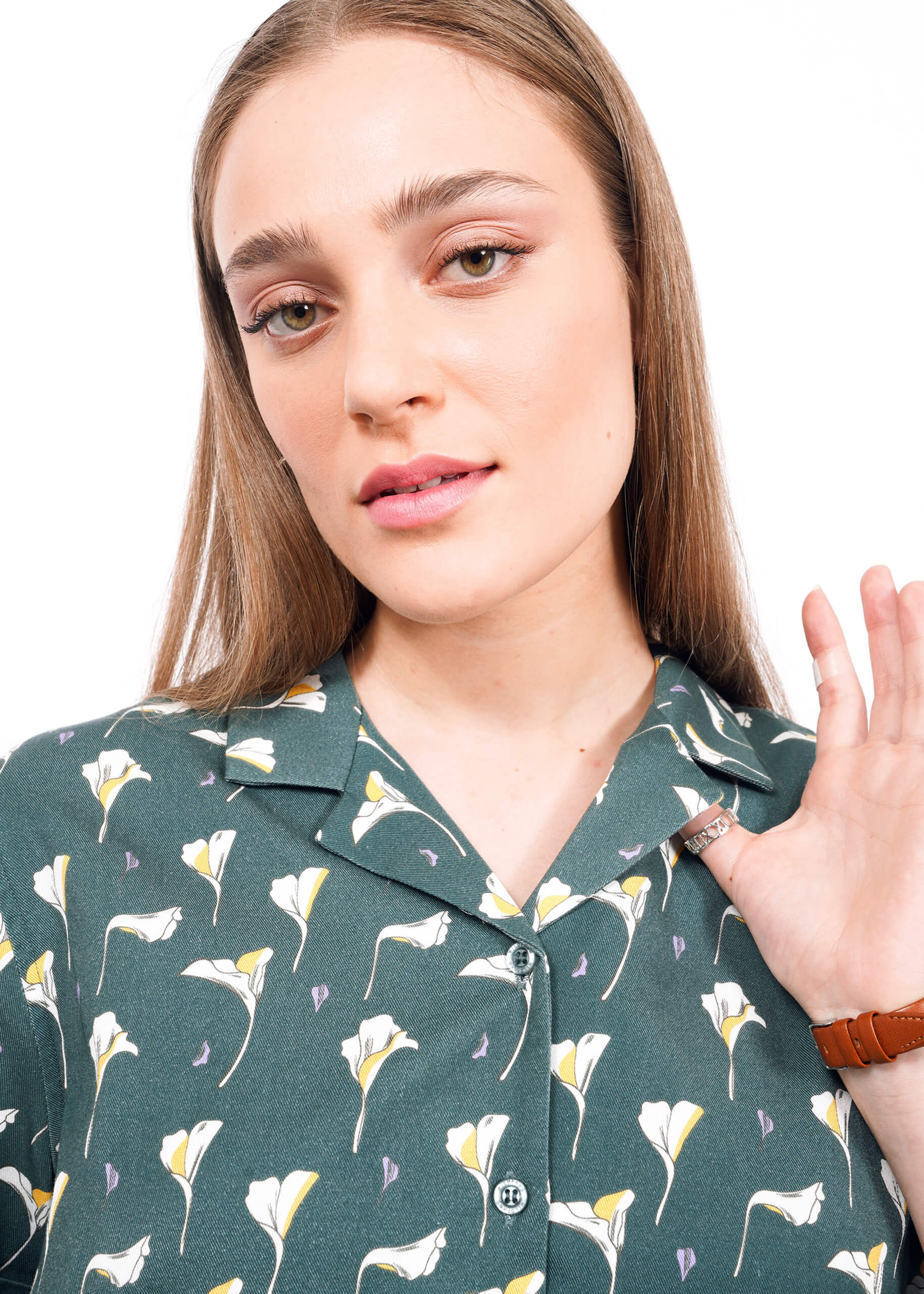 A young woman with straight, light brown hair wears The Everyday Camp Button Up—a green shirt with white flowers and a camp collar. She looks at the camera with a neutral expression, raising her left hand near her face.