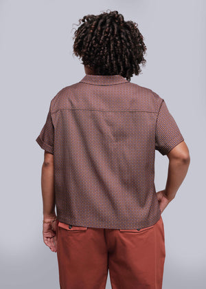 1X=Person wearing a patterned brown shirt and red pants against a plain background