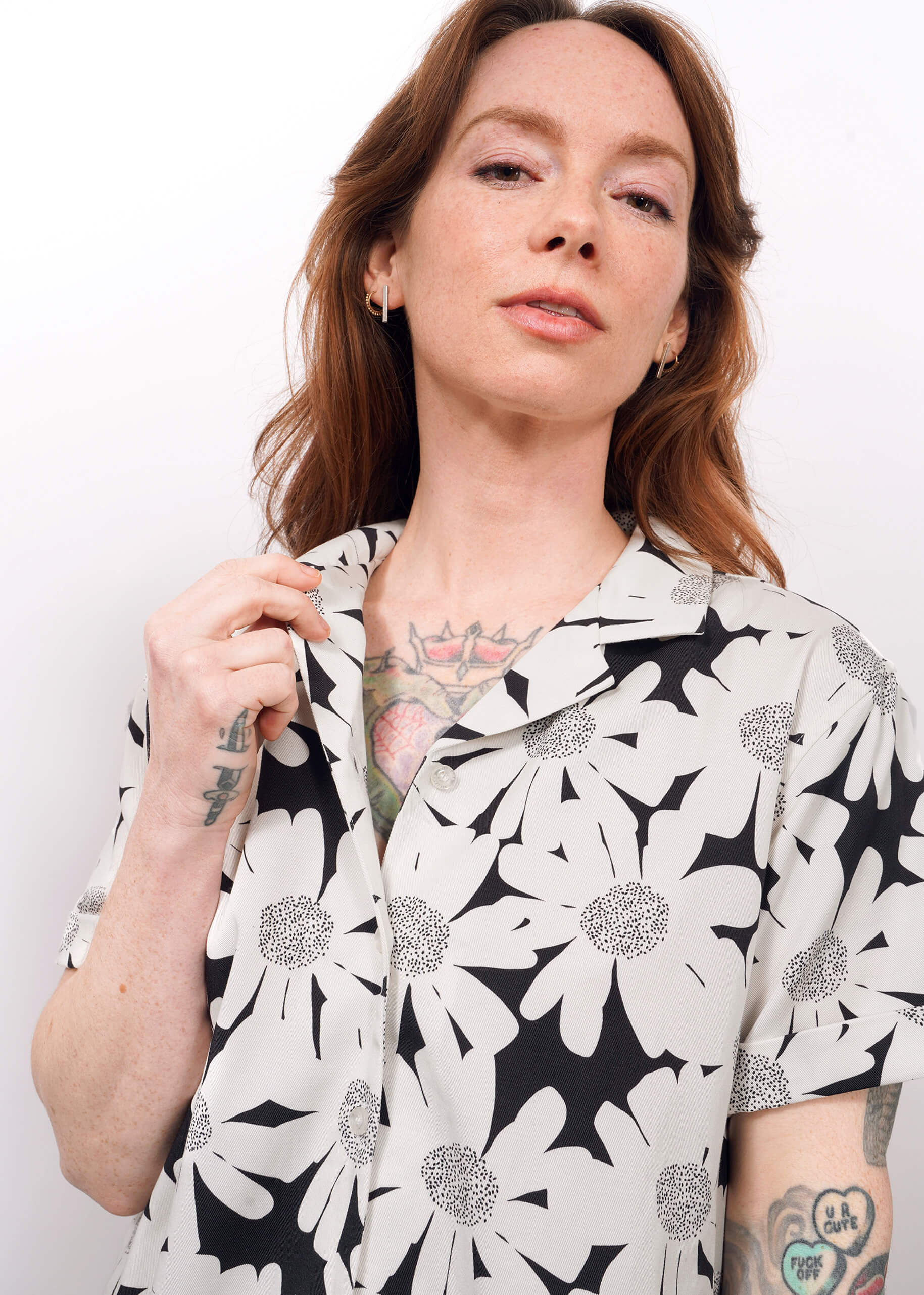 A woman with long reddish hair, freckles, and tattoos on her hand and chest wears The Everyday Camp Button Up—a black and white floral camp-collar shirt in relaxed Tencel—standing against a plain white background.
