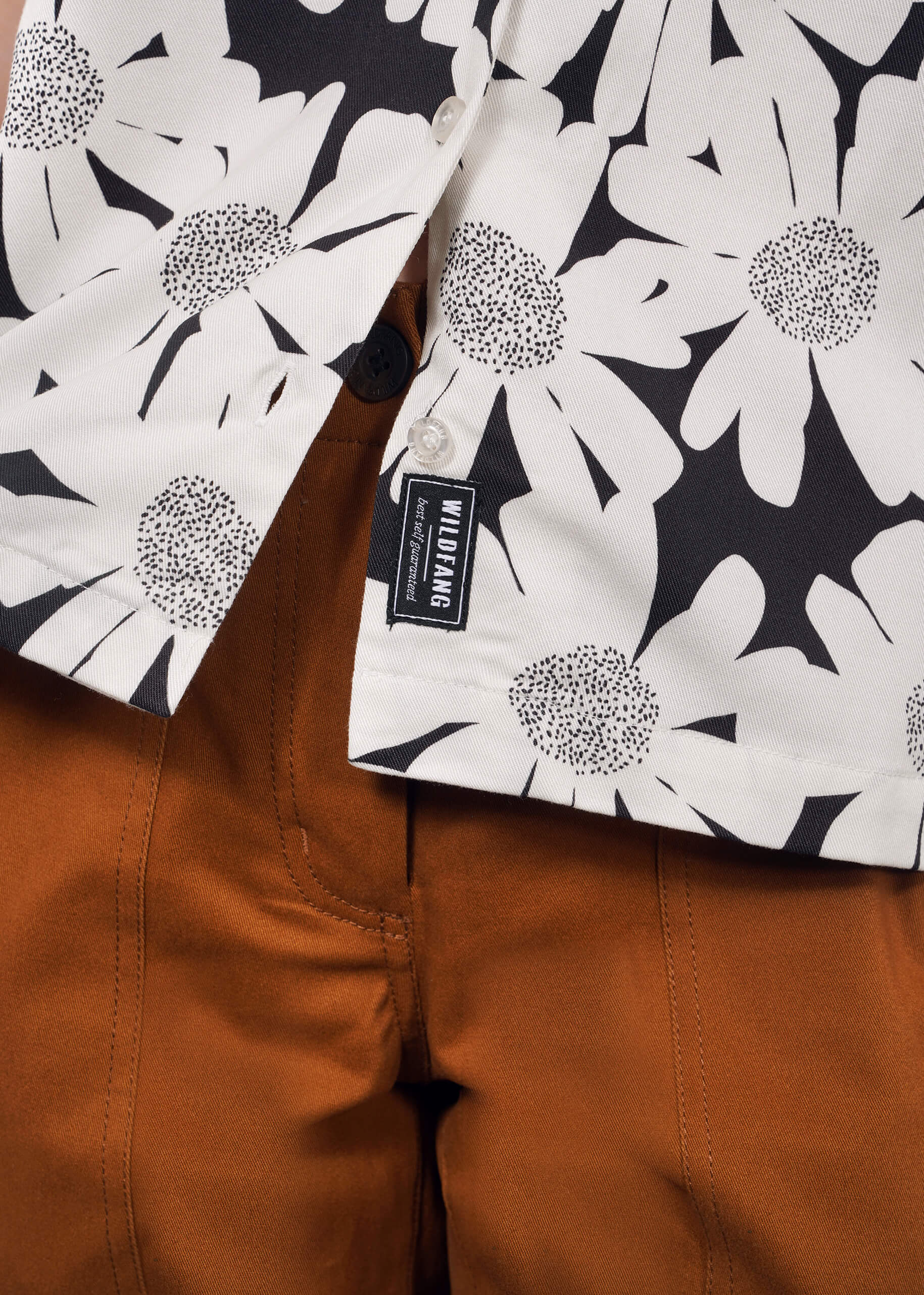 A close-up of a person wearing rust-colored pants and The Everyday Camp Button Up, a relaxed fit white shirt with large black dotted floral patterns and a black and white logo tag near the buttons.