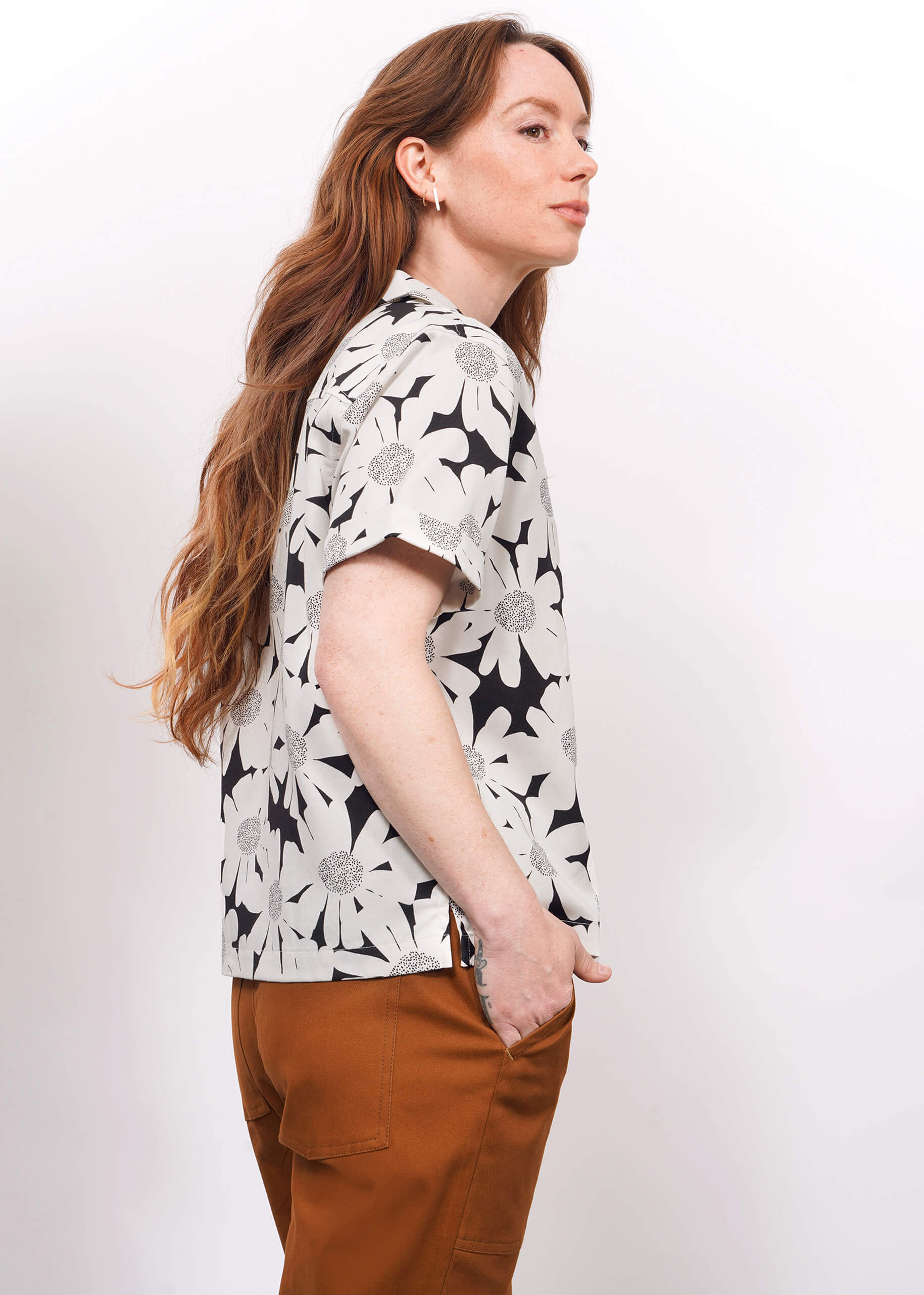 A person with long, wavy brown hair stands sideways against a white background, wearing The Everyday Camp Button Up—a relaxed fit black and white floral shirt in soft Tencel—and brown pants, with one hand in their pocket.