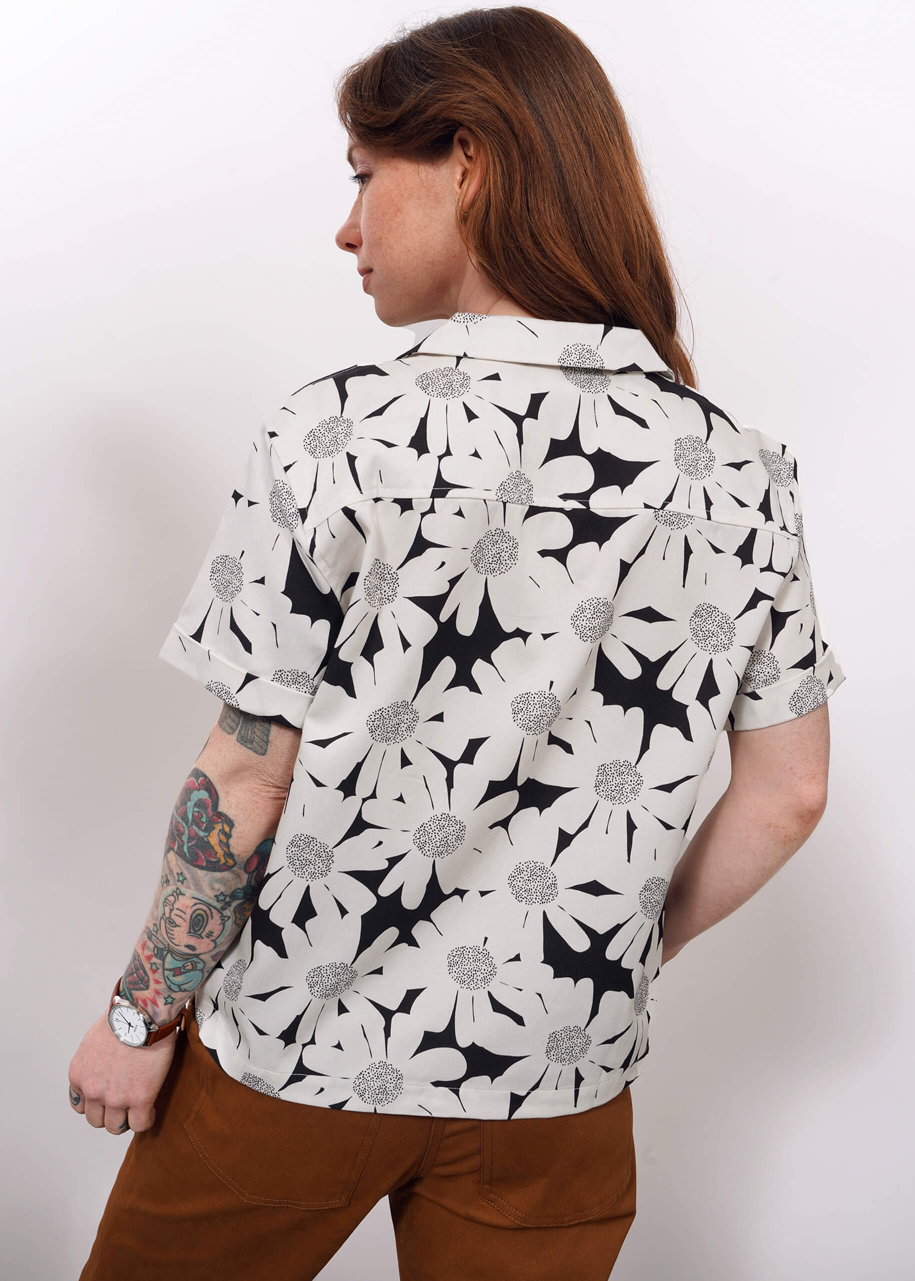 A person with long brown hair and visible arm tattoos is wearing The Everyday Camp Button Up in a relaxed floral print, standing with their back to the camera against a plain white background.