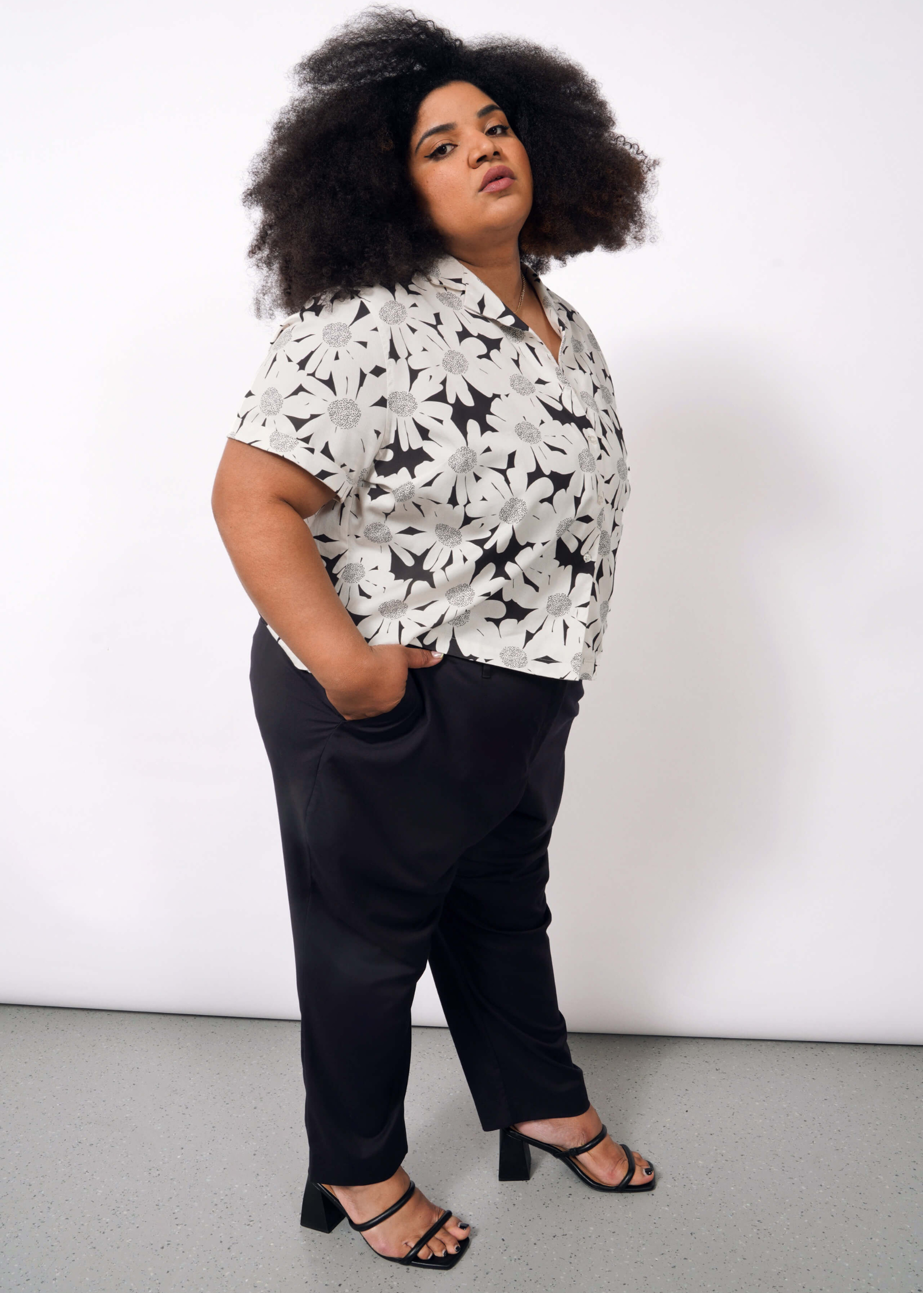 A person with curly hair stands confidently against a white background, wearing The Everyday Camp Button Up in a relaxed fit and floral pattern, paired with dark pants and black heeled sandals, hands in their pockets.