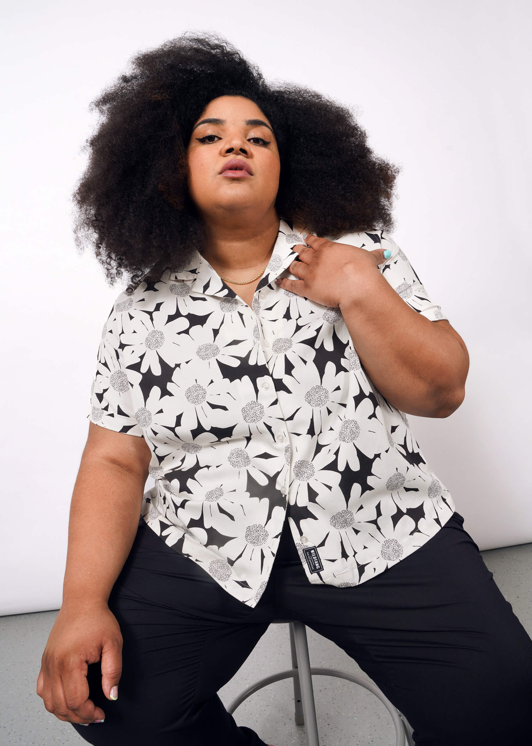 A person with voluminous curly hair sits on a stool wearing The Everyday Camp Button Up, a relaxed fit shirt with a bold white flower pattern, paired with black pants, posing confidently against a plain white background.