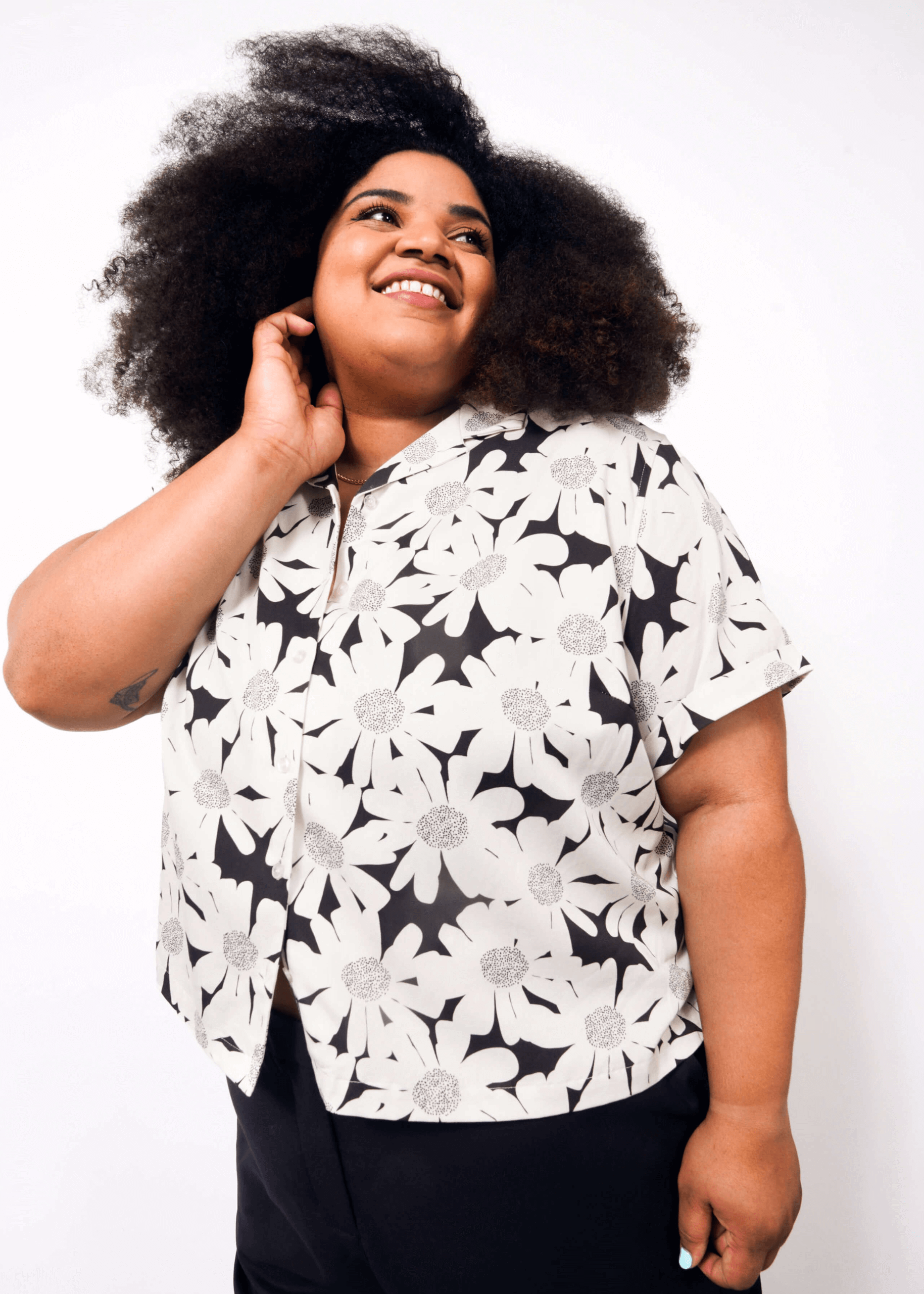 A smiling person with voluminous curly hair wears The Everyday Camp Button Up, a relaxed-fit black-and-white floral shirt, and dark pants while posing with one hand touching their neck against a white background.