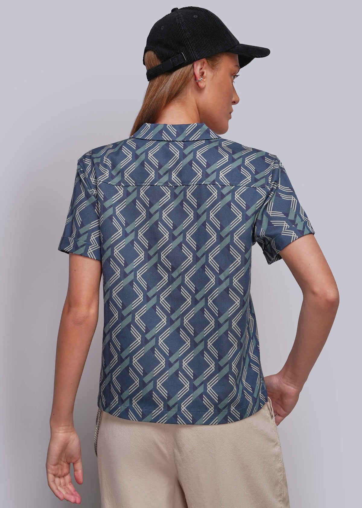 S=A man wearing a short-sleeve button-up shirt with a blue, gray, and white geometric pattern, paired with beige pants and a black baseball cap, shown from a back view against a light gray studio background.