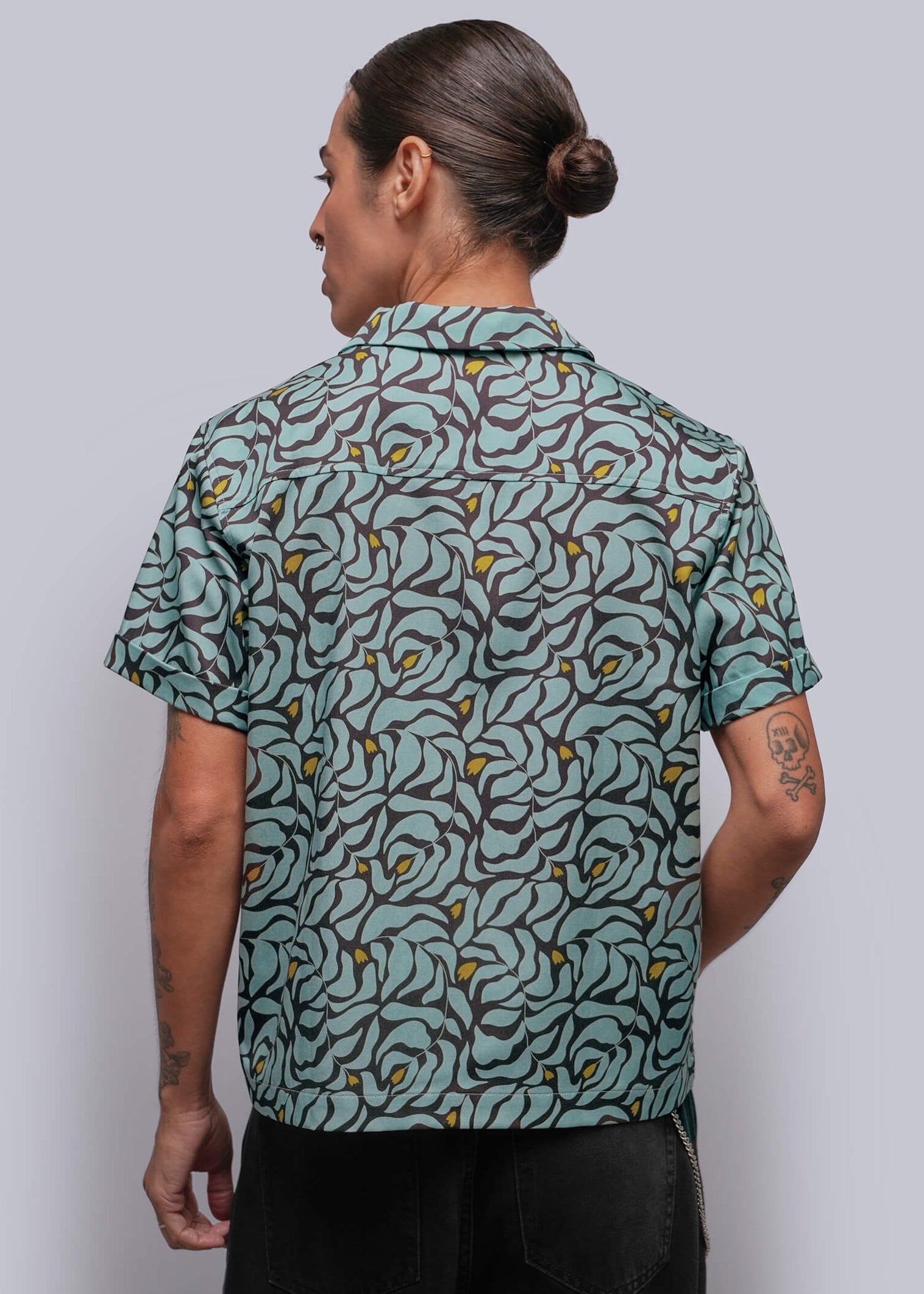 S=A person viewed from the back wearing a short-sleeve button-up shirt with a teal, gray, and yellow abstract floral pattern, paired with black pants, against a solid gray studio background.