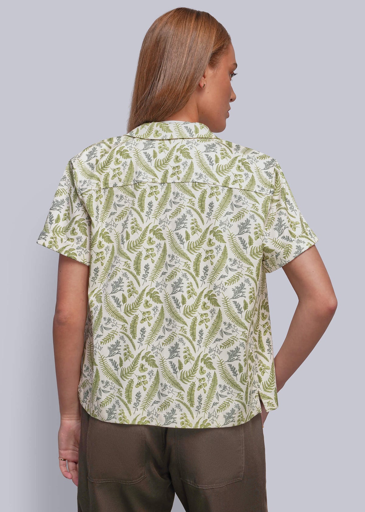 S=A woman shown from the back wearing a short-sleeve button-up shirt with an olive green and cream botanical pattern, paired with brown pants, against a solid gray studio background.