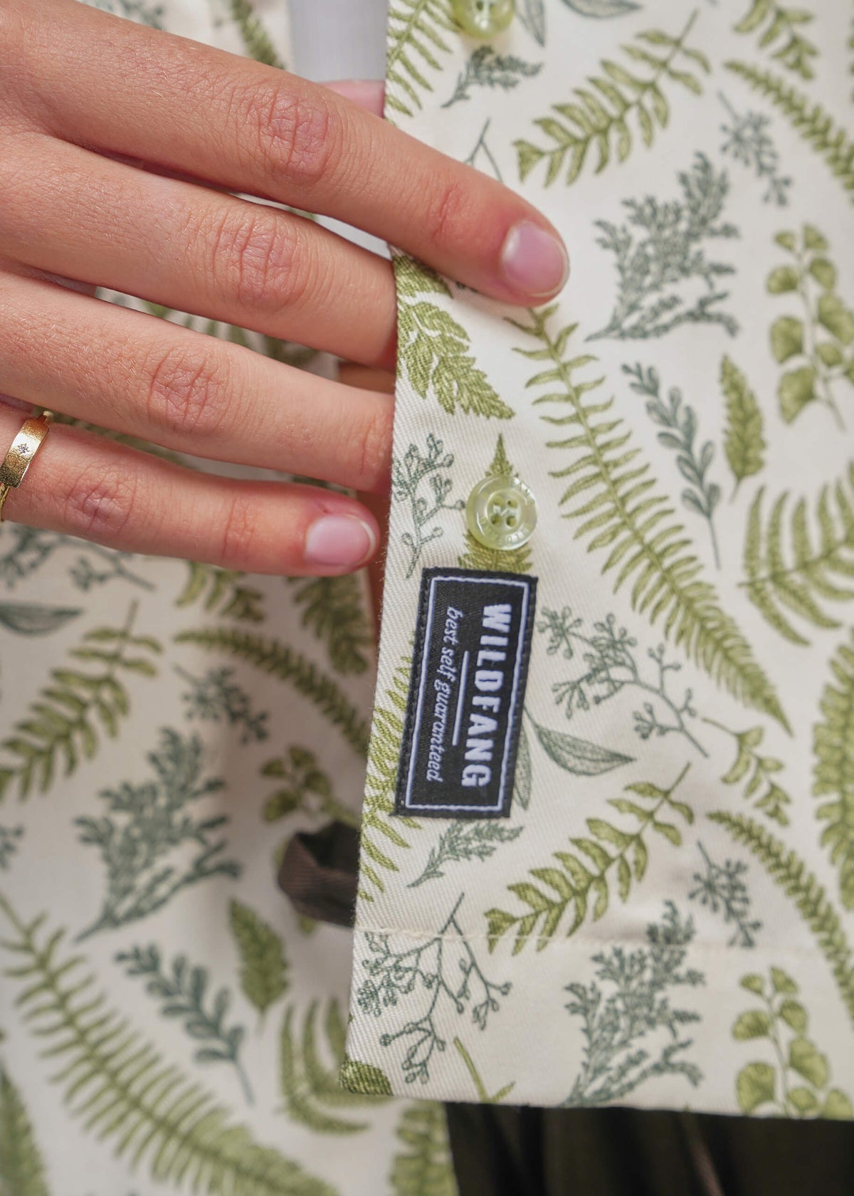 S=Close-up of a person's hand holding a buttoned flap of a shirt with a green fern botanical print on a cream background. A black brand label reading 'WILDFANG' is visible.