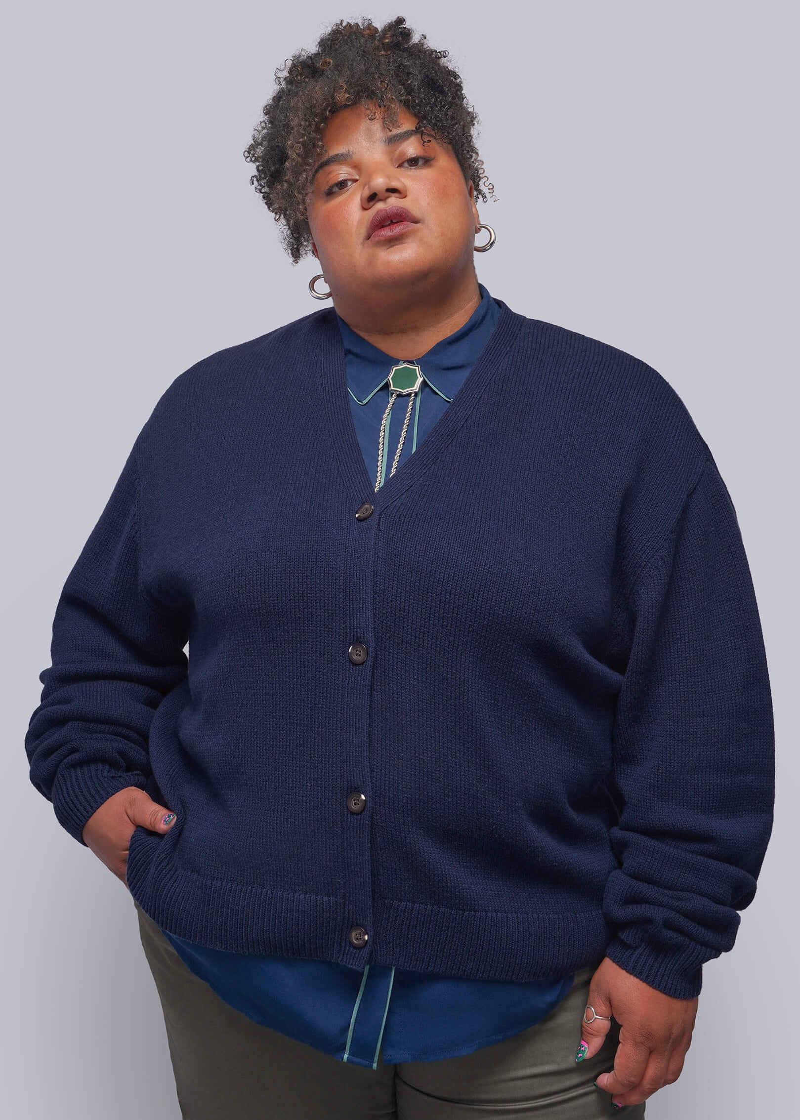 1X=A plus-size model wearing a navy blue knit cardigan over a blue collared shirt with a turquoise bolo tie, paired with olive green pants, against a solid gray studio background.
